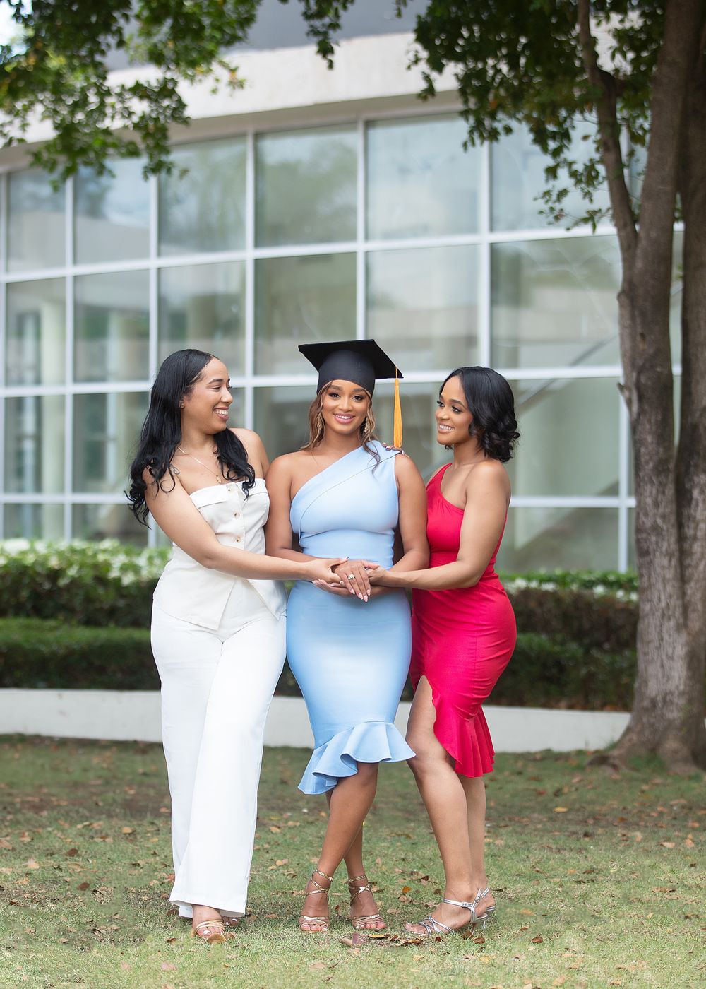 Three women celebrate a graduation outdoors, the graduate wearing a cap and light blue dress flanked by friends in white and red.