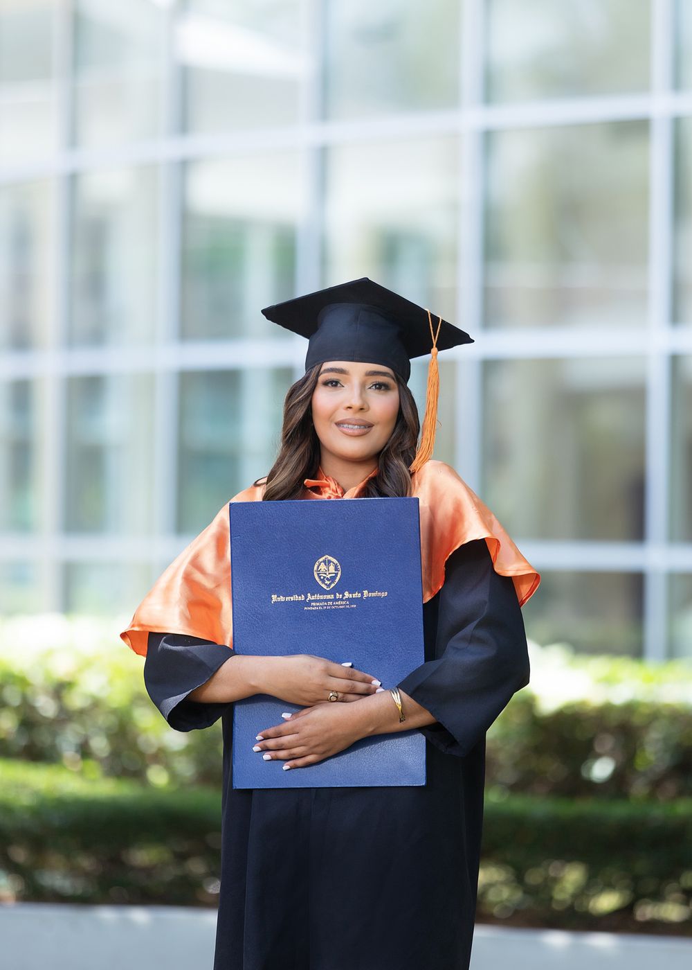 Woman in black graduation gown with orange hood holding diploma, wearing mortarboard cap outdoors.