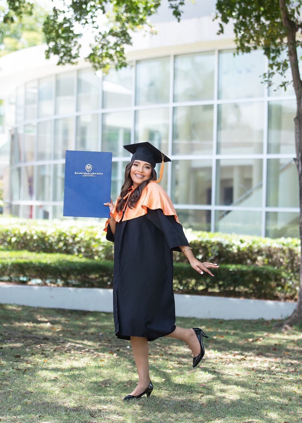 Smiling female graduate in black and orange cap and gown holding diploma outdoors on campus lawn.