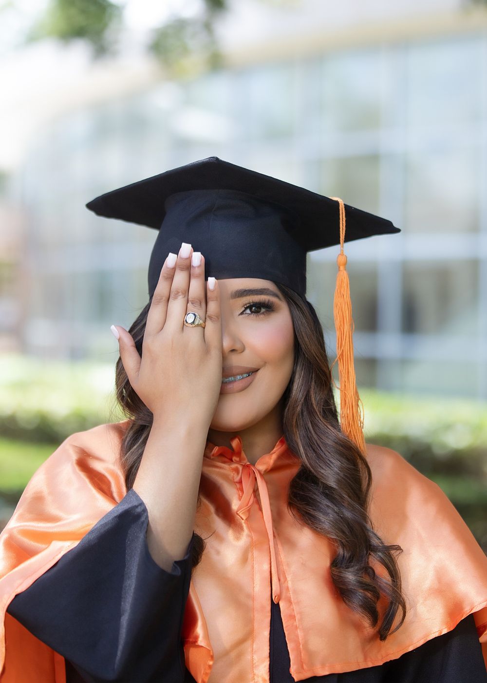 Young woman in orange and black graduation cap and gown covering one eye with her hand, smiling outdoors.