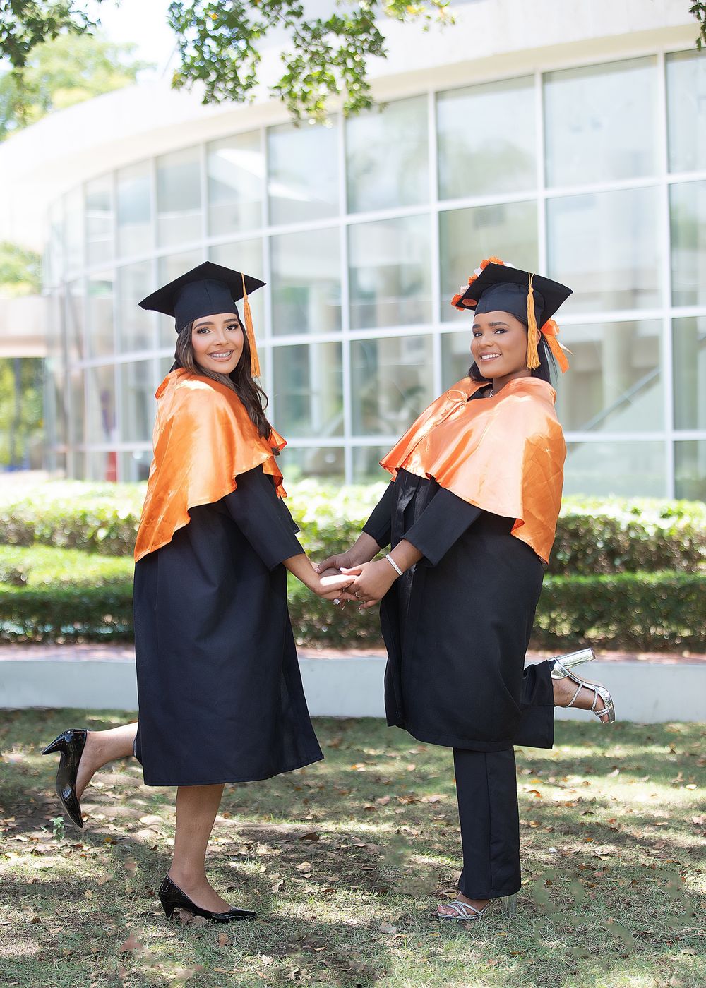 Two women in black and orange graduation caps and gowns pose playfully outdoors on a sunny day.