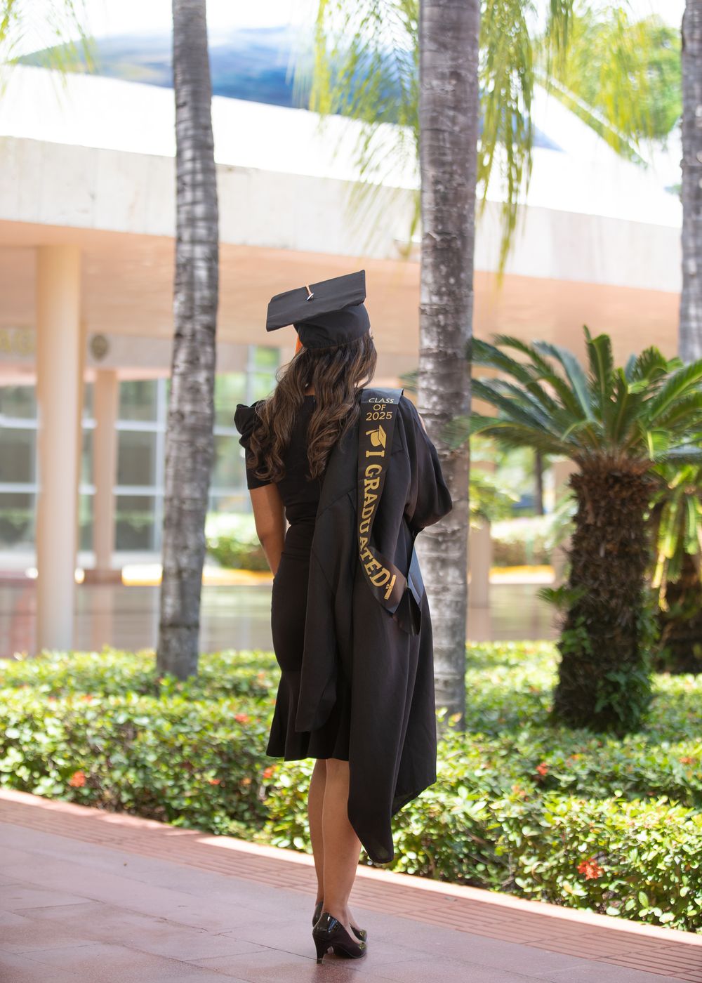 Graduate in black cap and gown with gold sash stands on campus walkway surrounded by palm trees.