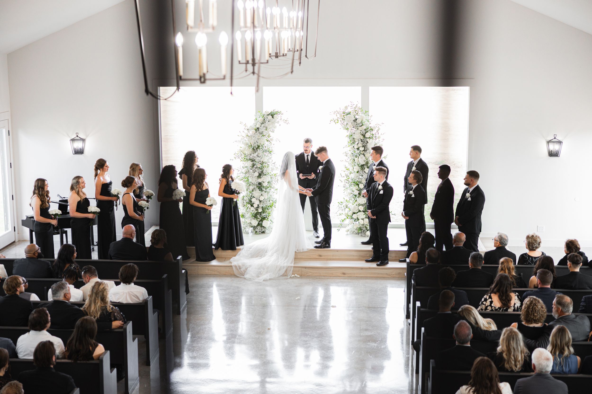 Elegant wedding ceremony in modern white venue with floral arch and guests in black attire.