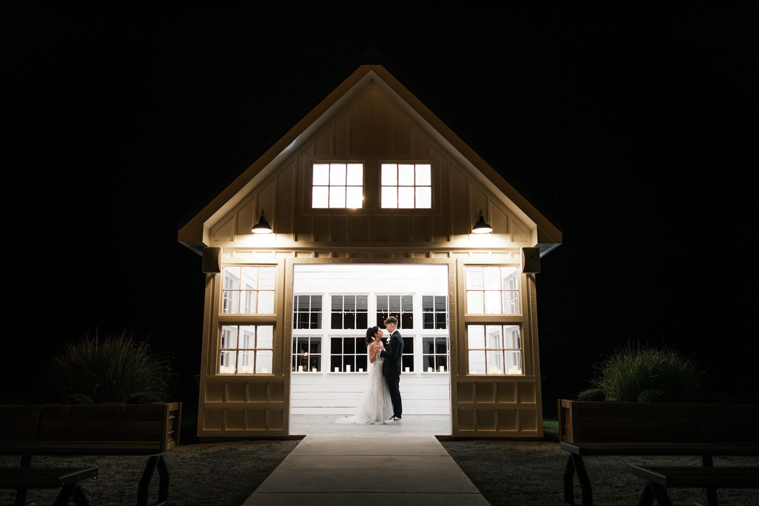 Newlyweds share a romantic kiss framed by illuminated barn doors against a dark night sky.