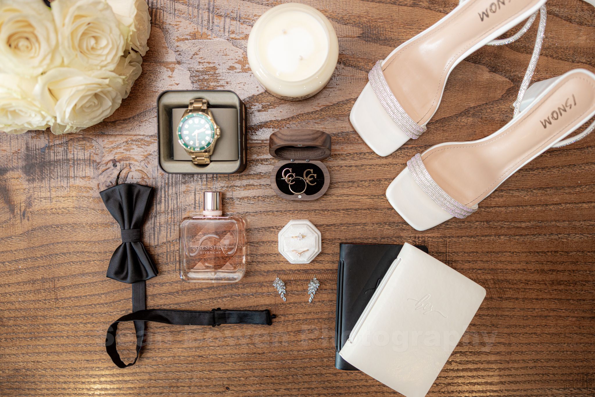 Wedding flat lay featuring white heels, perfume, watch, bow tie, rings, and white roses.