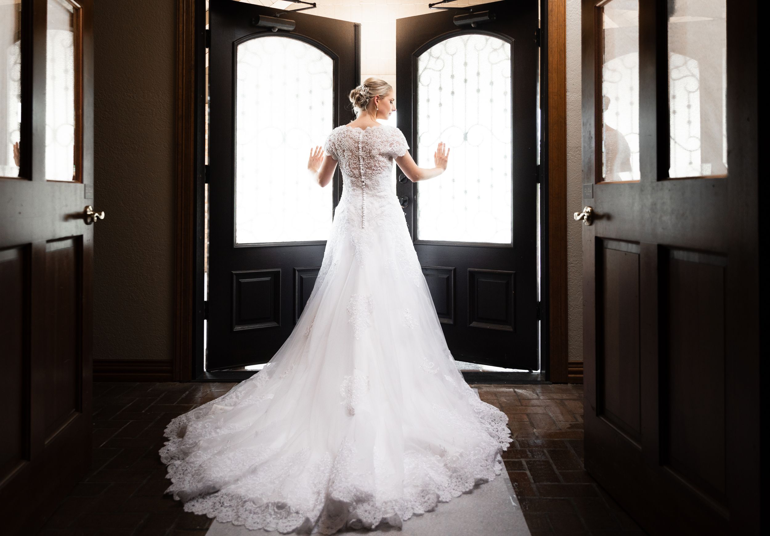 Bride in lace gown with cathedral train stands before elegant arched double doors.