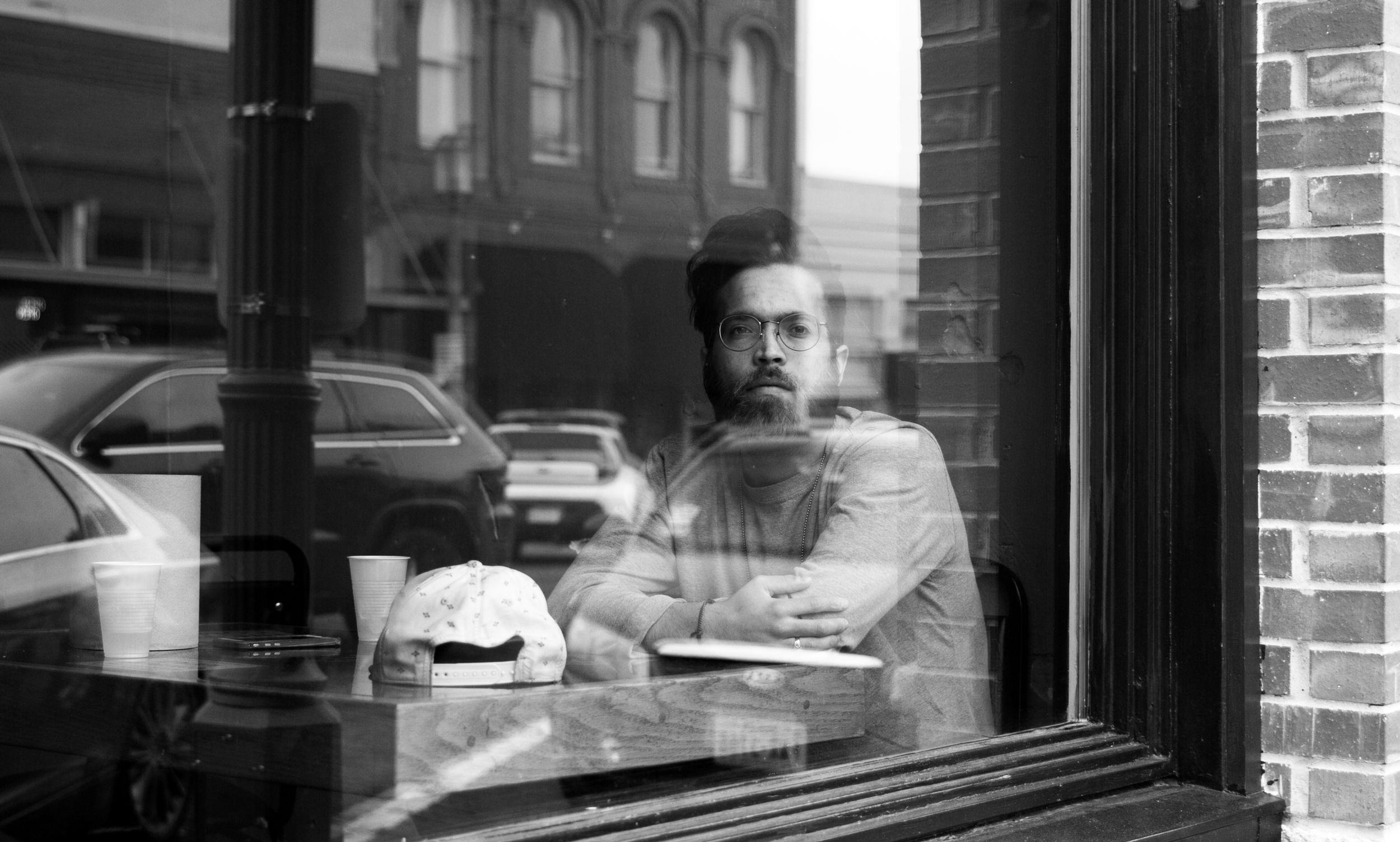 Black and white portrait of bearded man sitting alone in coffee shop window.