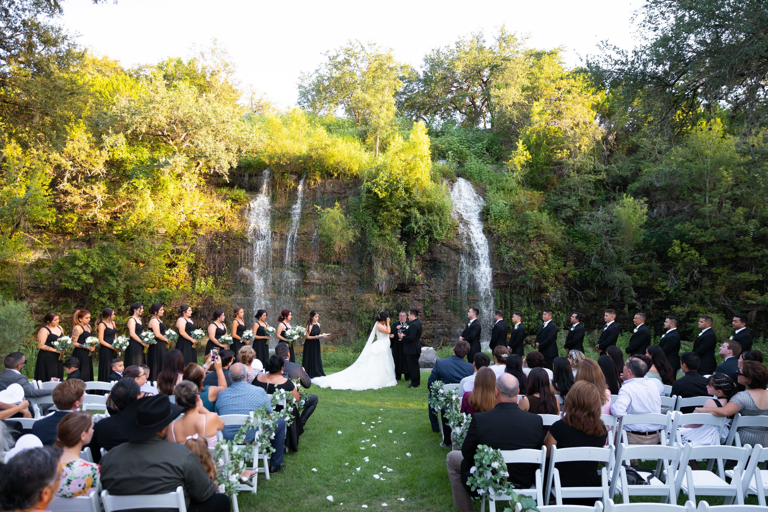 Outdoor wedding ceremony before stunning waterfall backdrop with guests seated on white chairs.