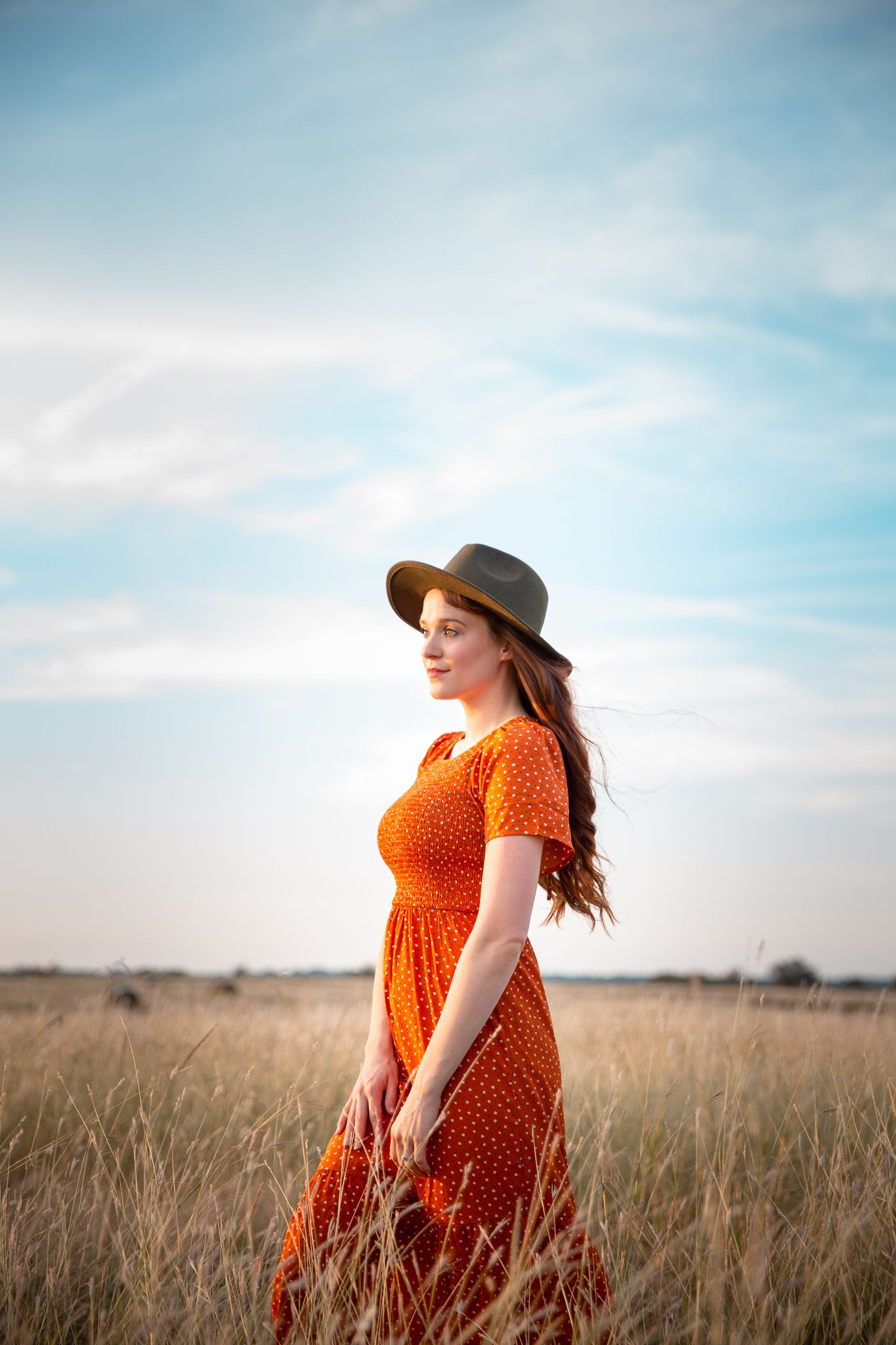 Woman in red polka dot dress and hat standing in golden wheat field under blue sky.