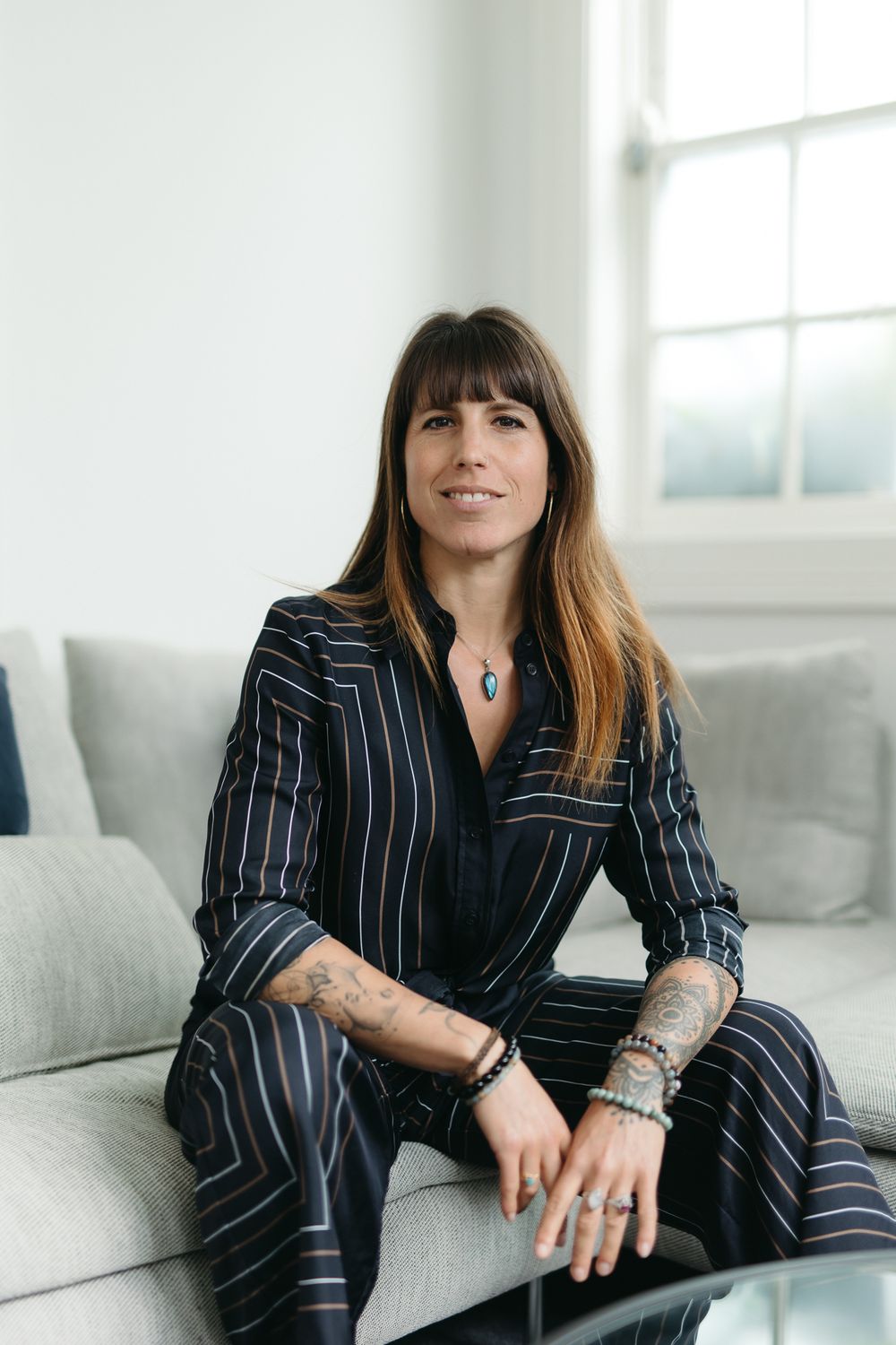 Woman in striped pantsuit with tattoos sits on gray sofa in bright modern room, smiling confidently.