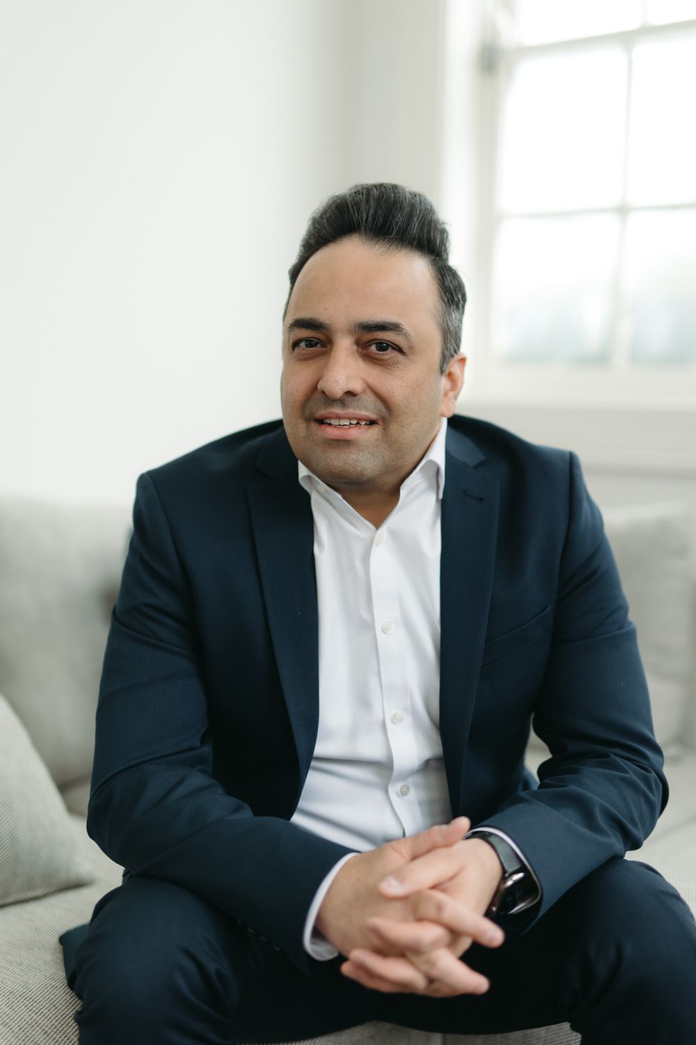 A professional man in a navy suit sits on a sofa, smiling confidently with hands clasped, in a bright modern room.