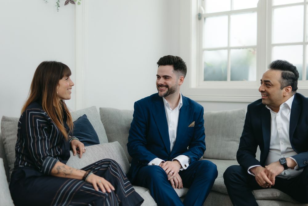 Three professionals in business attire smiling and conversing on a gray sofa in a bright modern room.