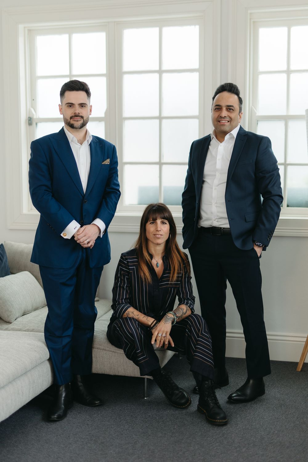 Three professional real estate agents posing in a bright modern office, two men in blue suits and a woman in striped outfit.