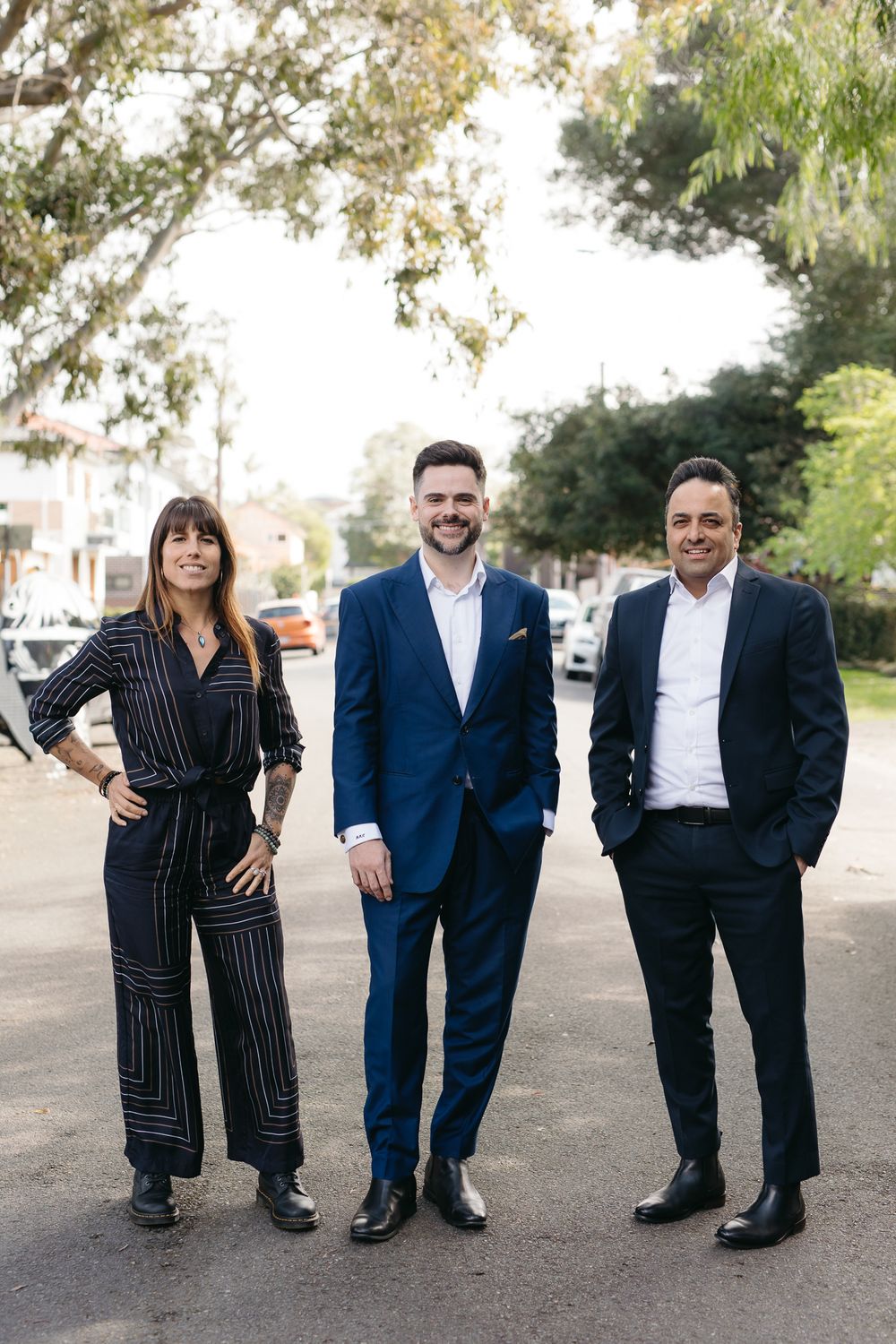 Three real estate professionals in formal attire posing outdoors on a tree-lined street.