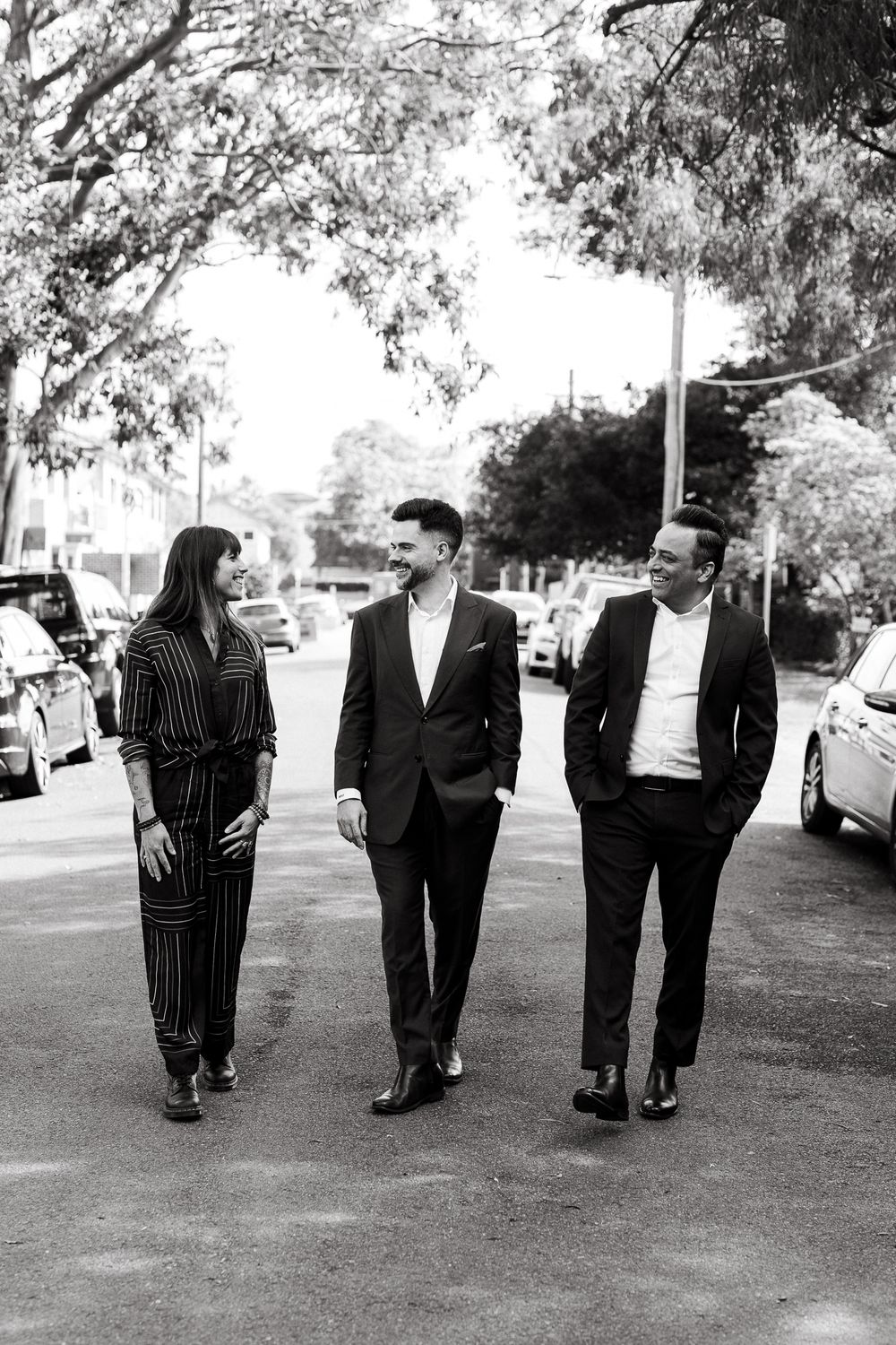 Three professionals in formal attire walking together on a tree-lined street in black and white photo.
