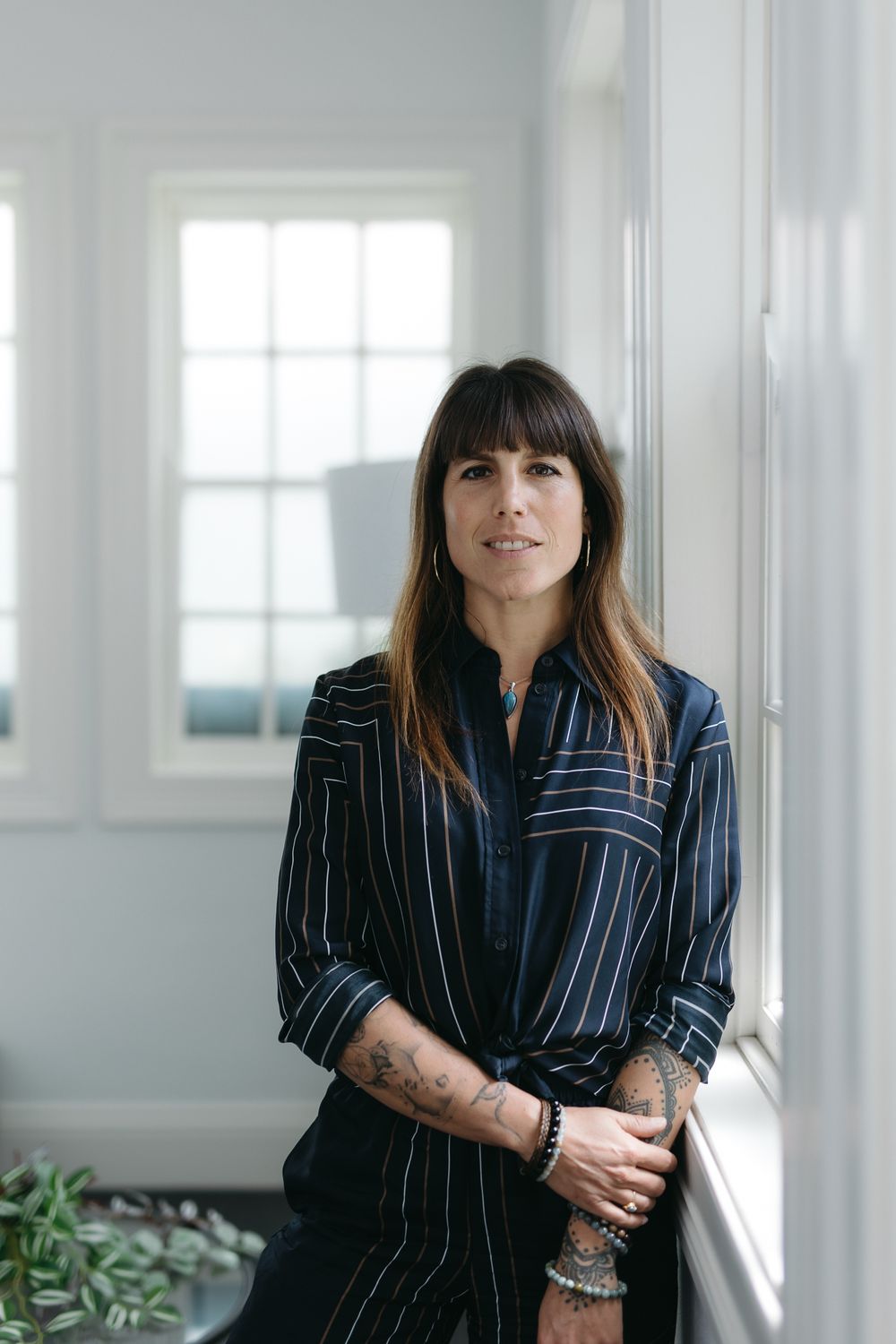 Woman in striped navy shirt leaning against white window frame with tattoos and layered bracelets.