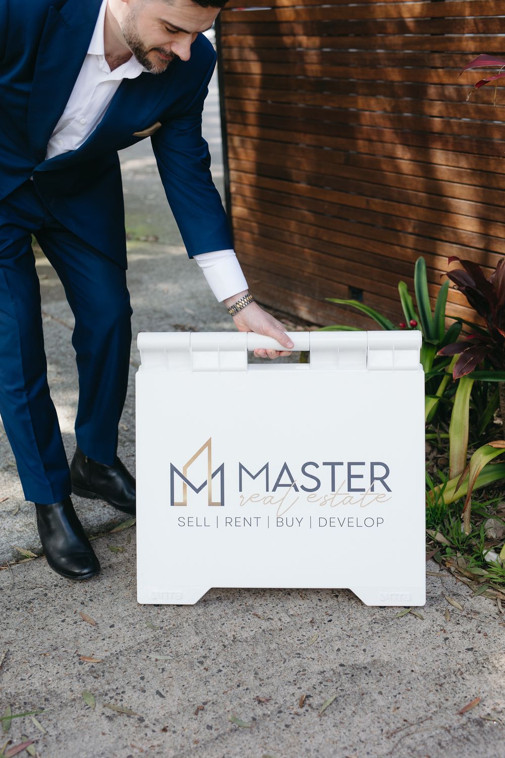 Man in blue suit placing a Master Real Estate sign reading Sell Rent Buy Develop outdoors.