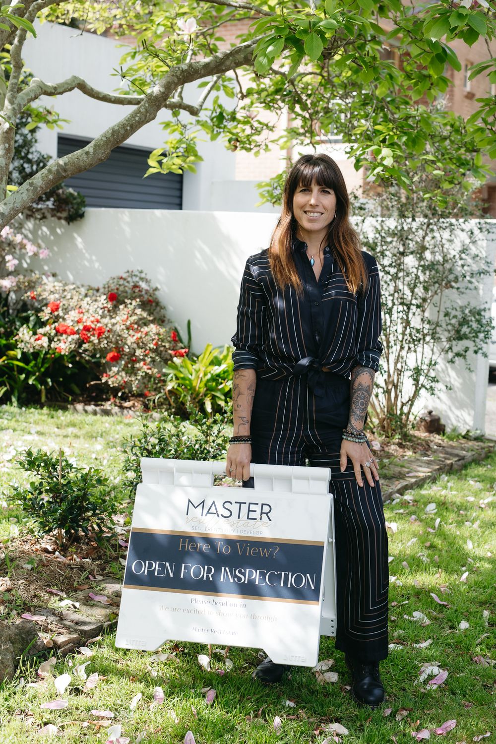 Woman in striped outfit holding a Master Real Estate open for inspection sign outdoors.