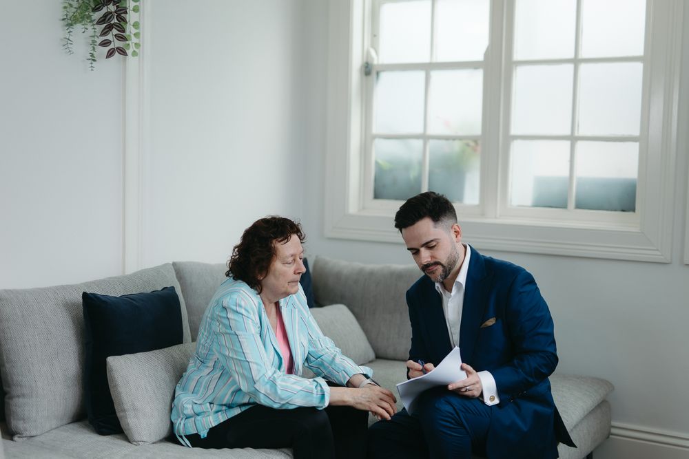 A financial advisor in a navy suit reviews documents with an older woman on a gray sofa in a bright living room.