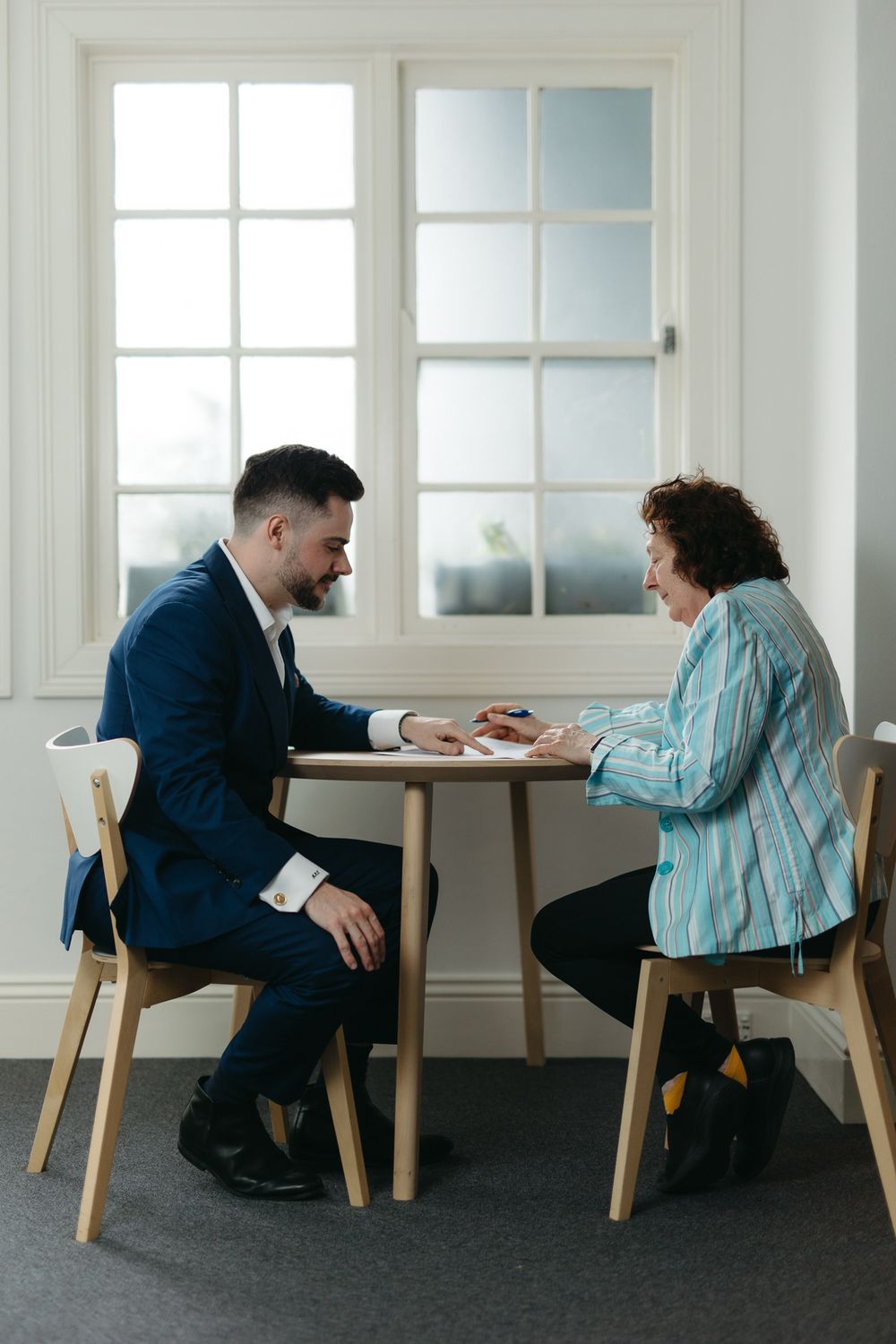 A businessman in a navy suit meets with a woman in a striped shirt at a small wooden table near a bright window.