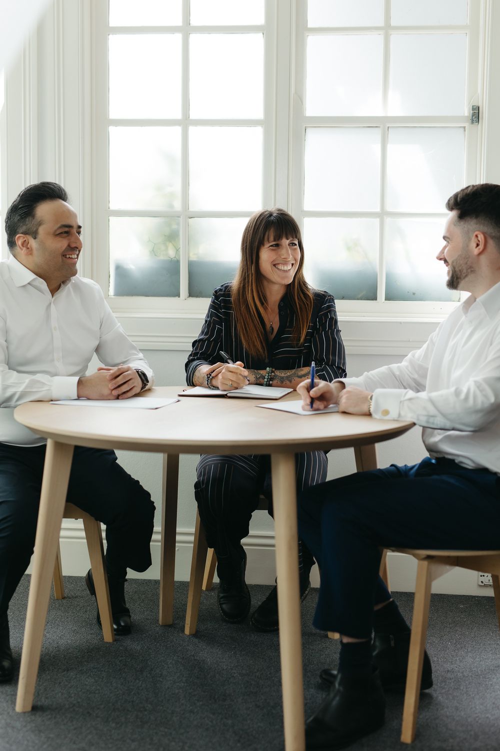 Three business professionals smiling and collaborating at a round wooden table in a bright office meeting room.