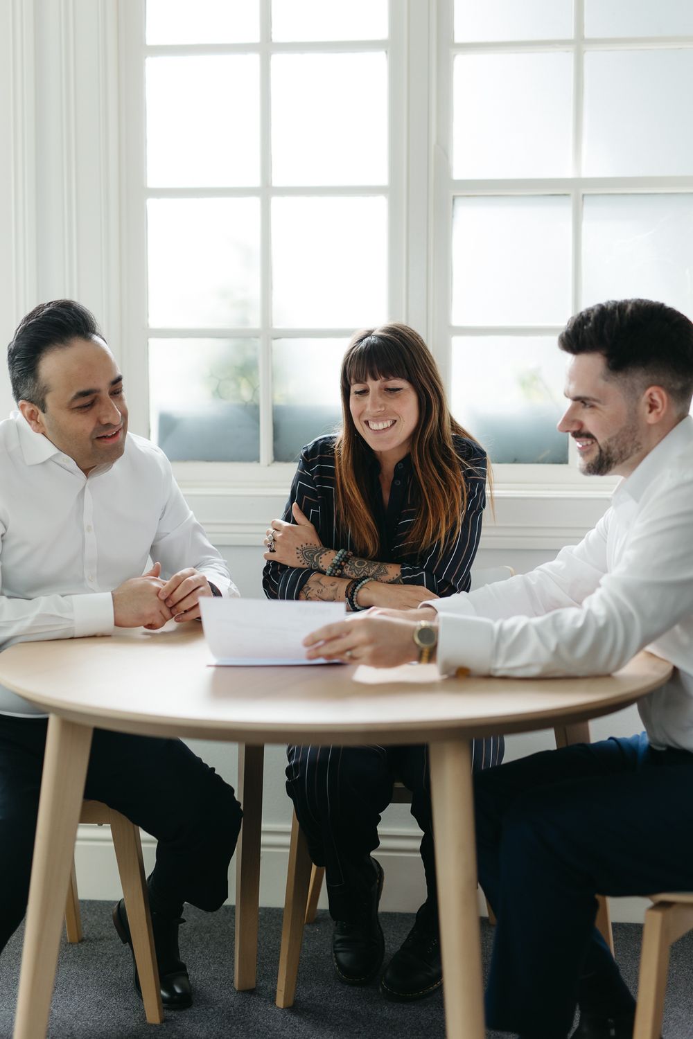 Three business professionals smiling and reviewing documents at a round wooden table in a bright modern office.