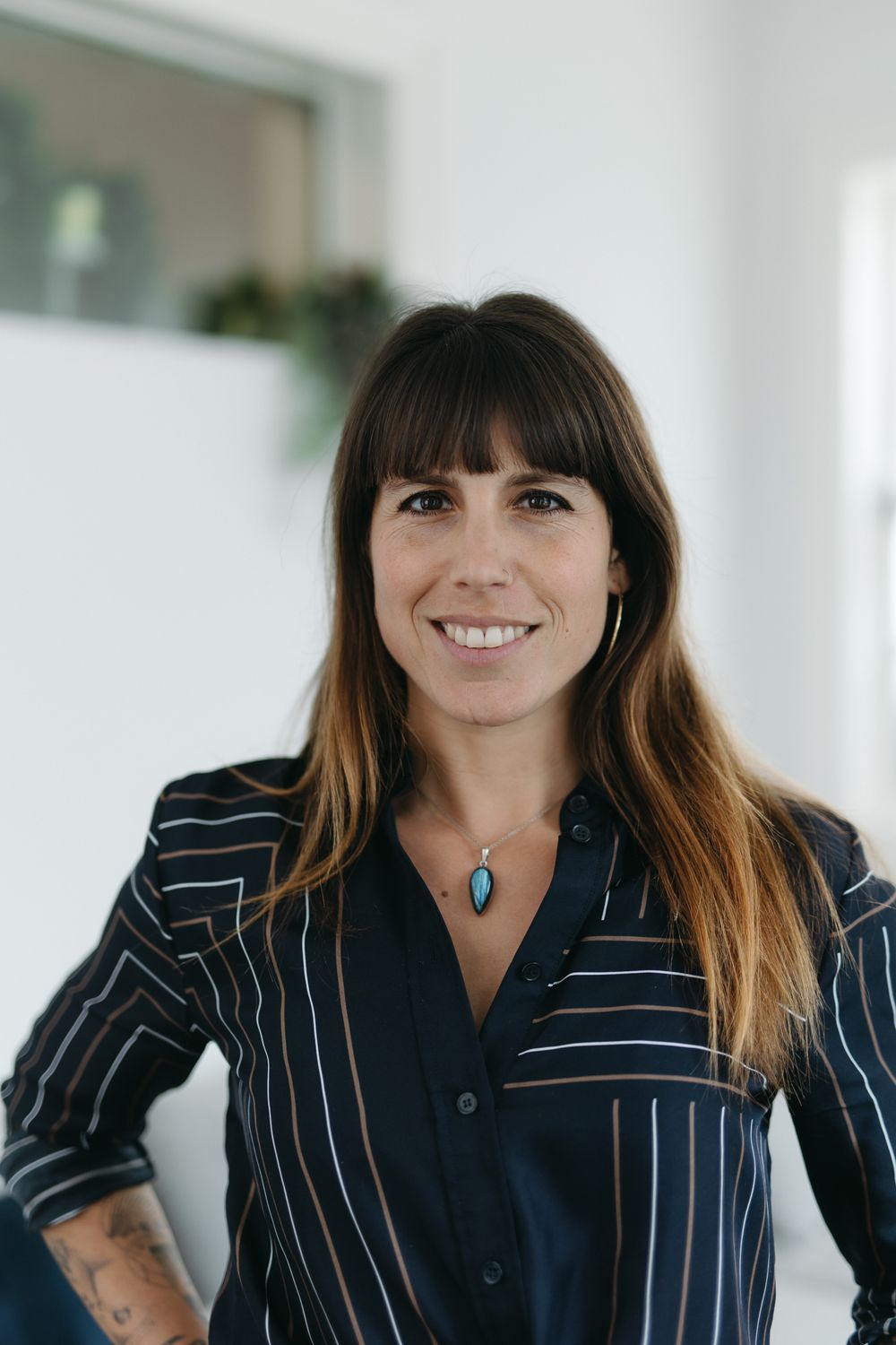 Smiling woman with long brown hair wearing a striped shirt and turquoise pendant necklace standing indoors.