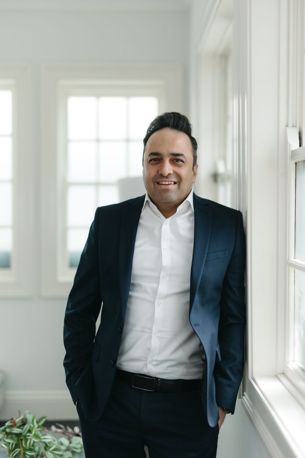 A smiling man in a navy suit and white shirt leans against a wall by bright windows in a modern interior.