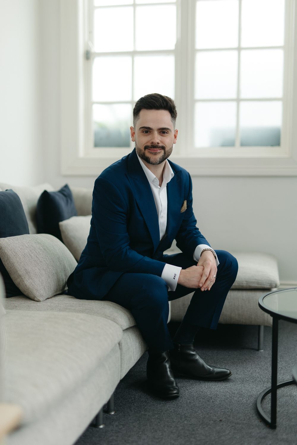 A well-dressed man in a navy blue suit sits confidently on a gray sofa, smiling in a bright modern interior.