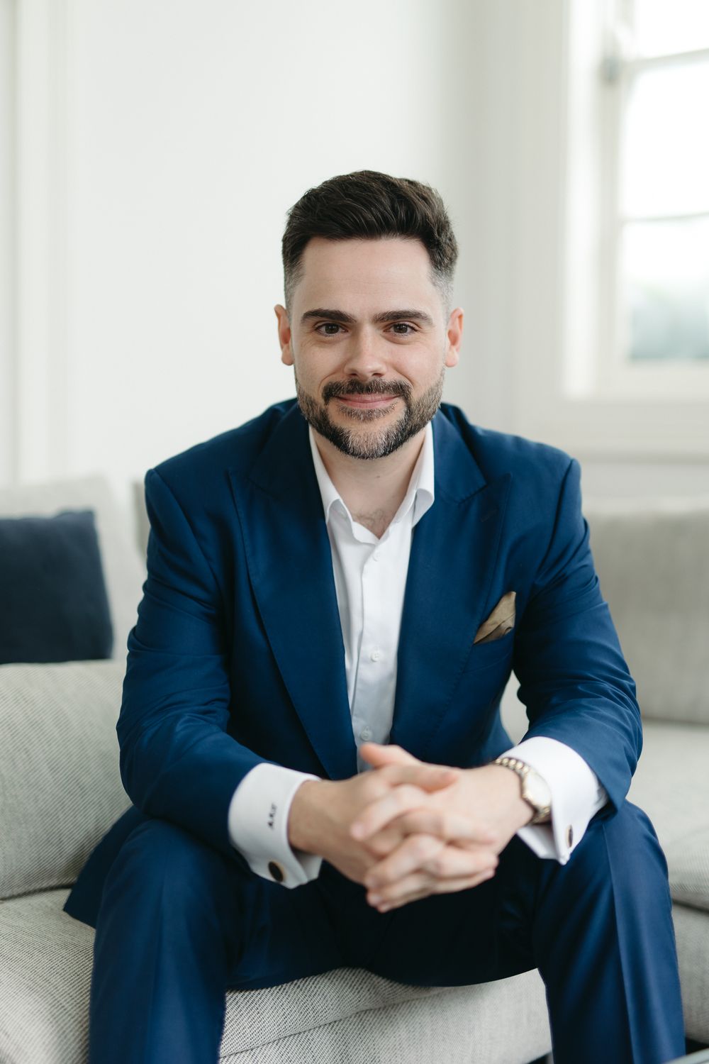 A smiling businessman in a navy blue suit sits confidently on a sofa with hands clasped.