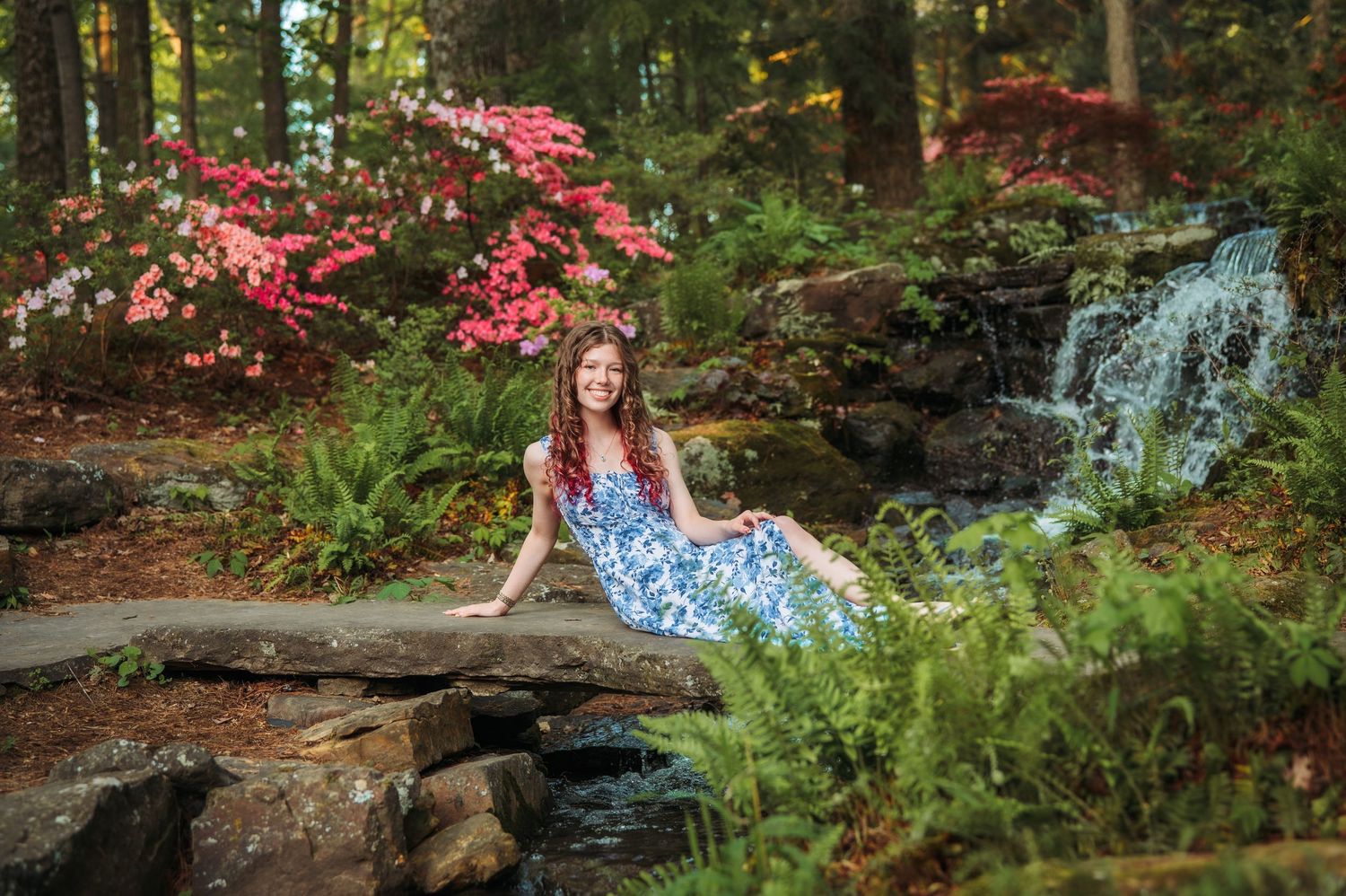 Young woman in blue floral dress sitting on rocks beside a waterfall in a lush garden with pink blooming trees.
