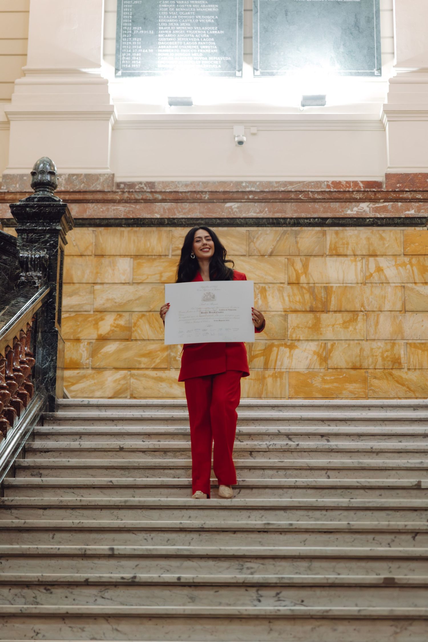 Woman in red suit stands on grand staircase holding a white diploma certificate.