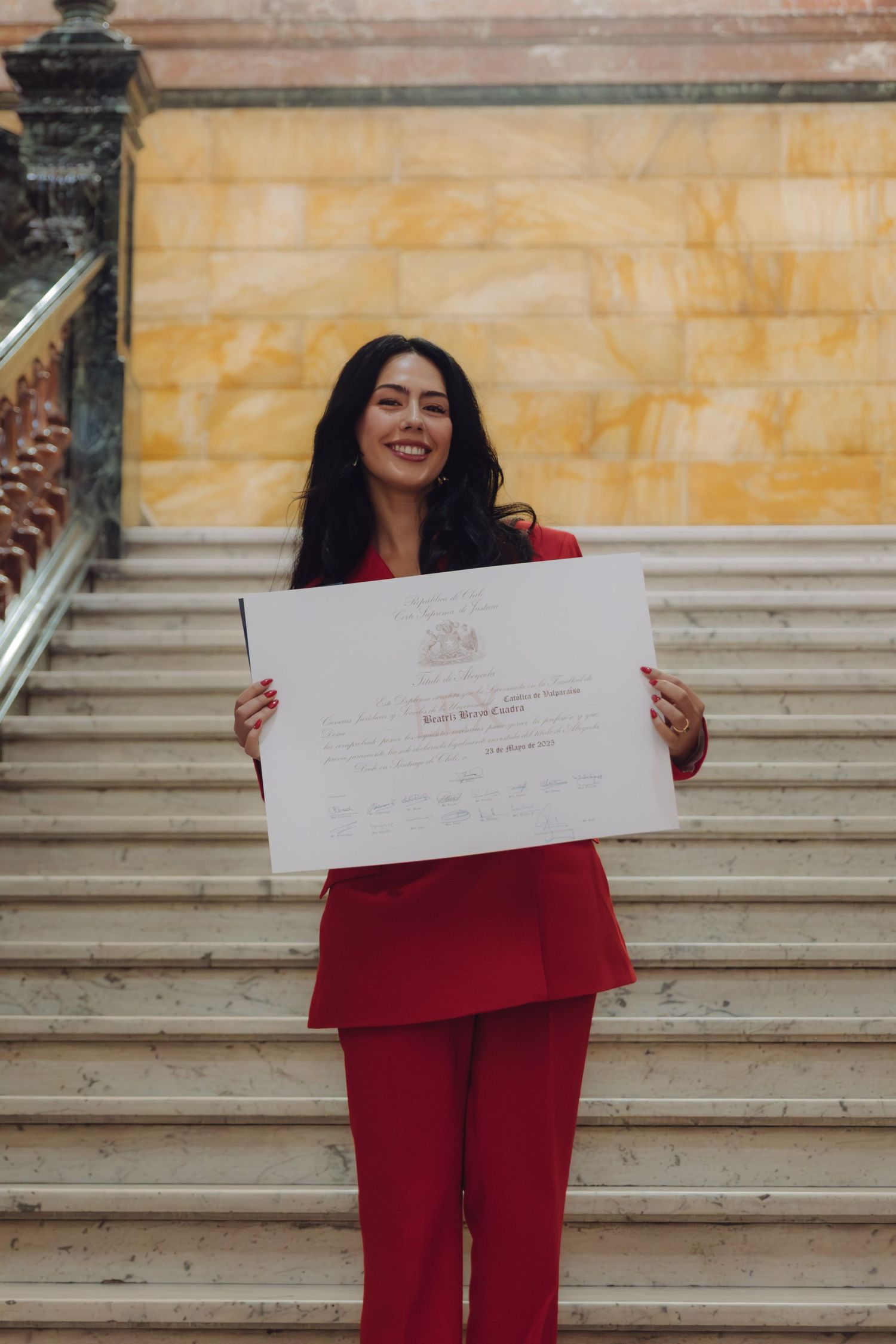 Woman in red suit smiles on marble stairs holding a large graduation diploma certificate.