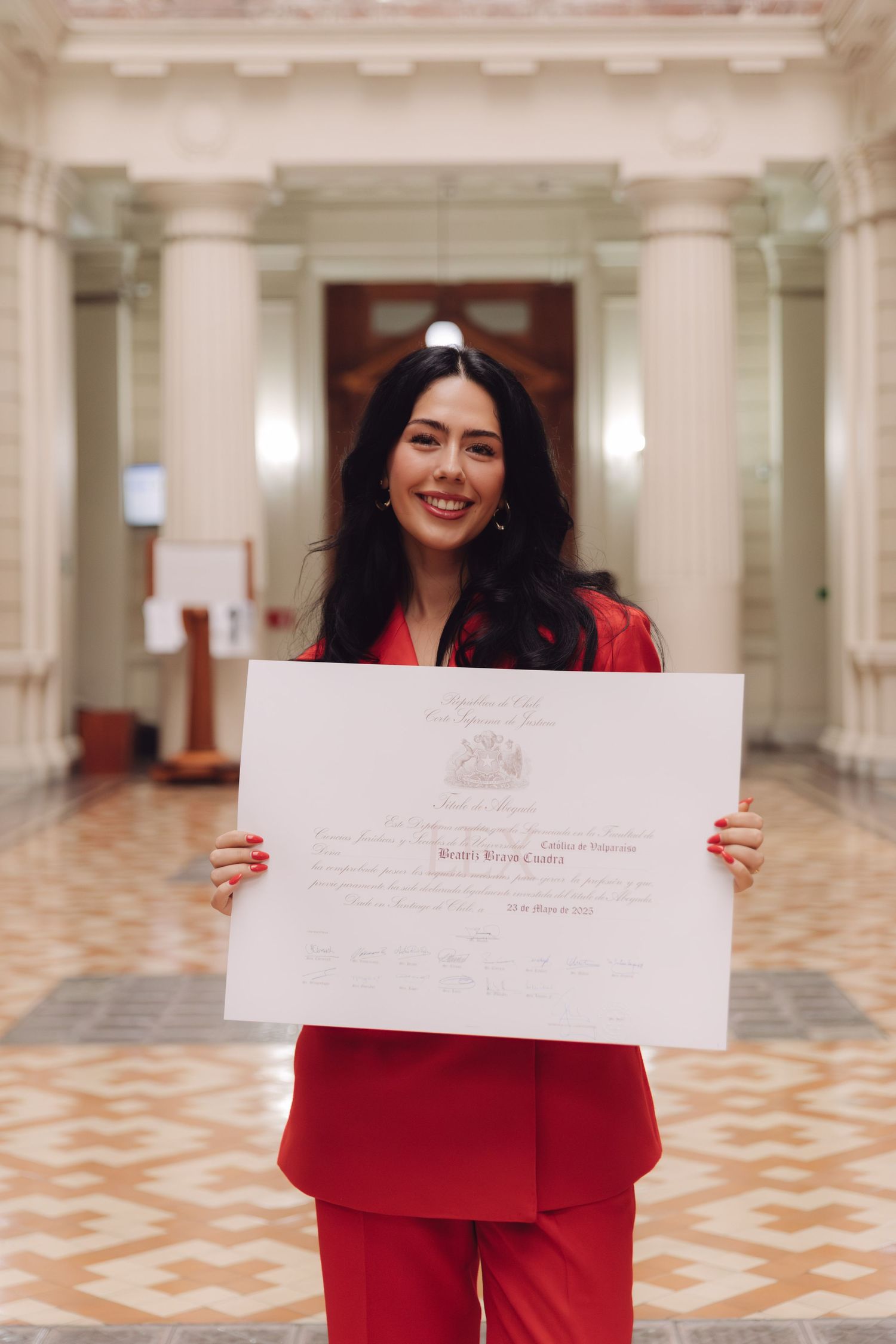 Smiling woman in red suit holds graduation diploma inside an elegant columned hall.