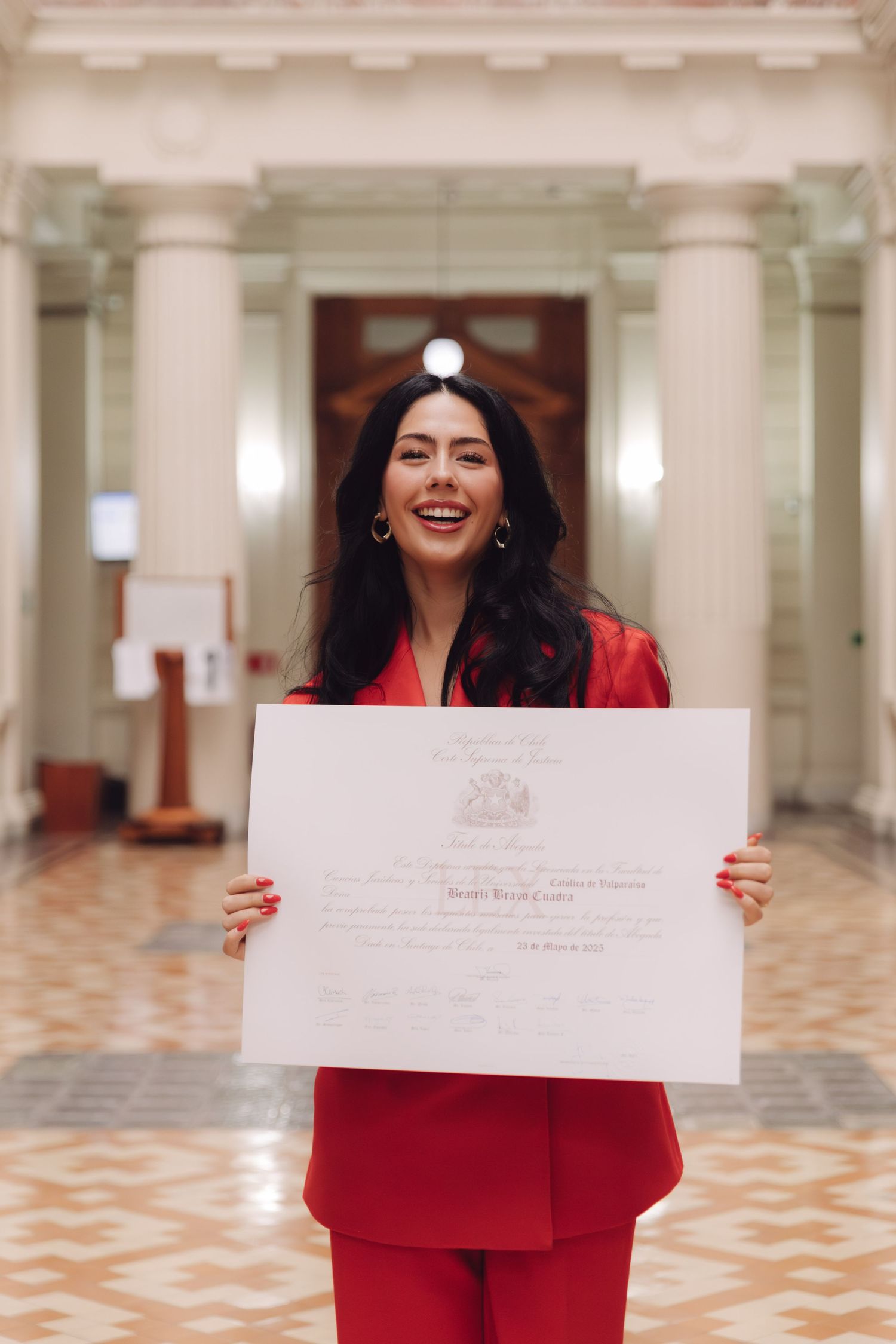 Joyful woman in red pantsuit proudly displays her graduation diploma in a grand interior hall.