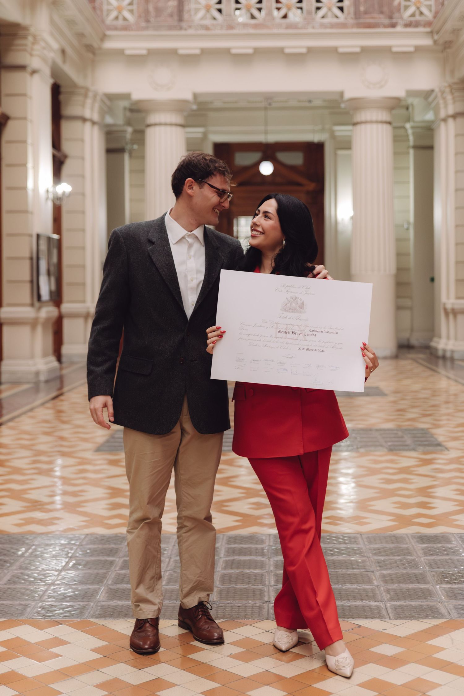 Couple smiles together inside elegant columned hall, woman in red holding a graduation diploma.