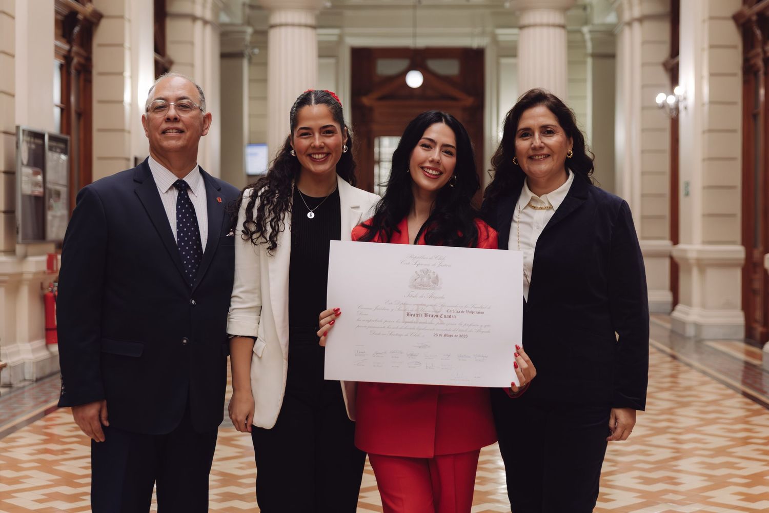 Graduate in red suit poses with three family members holding diploma in ornate hallway.