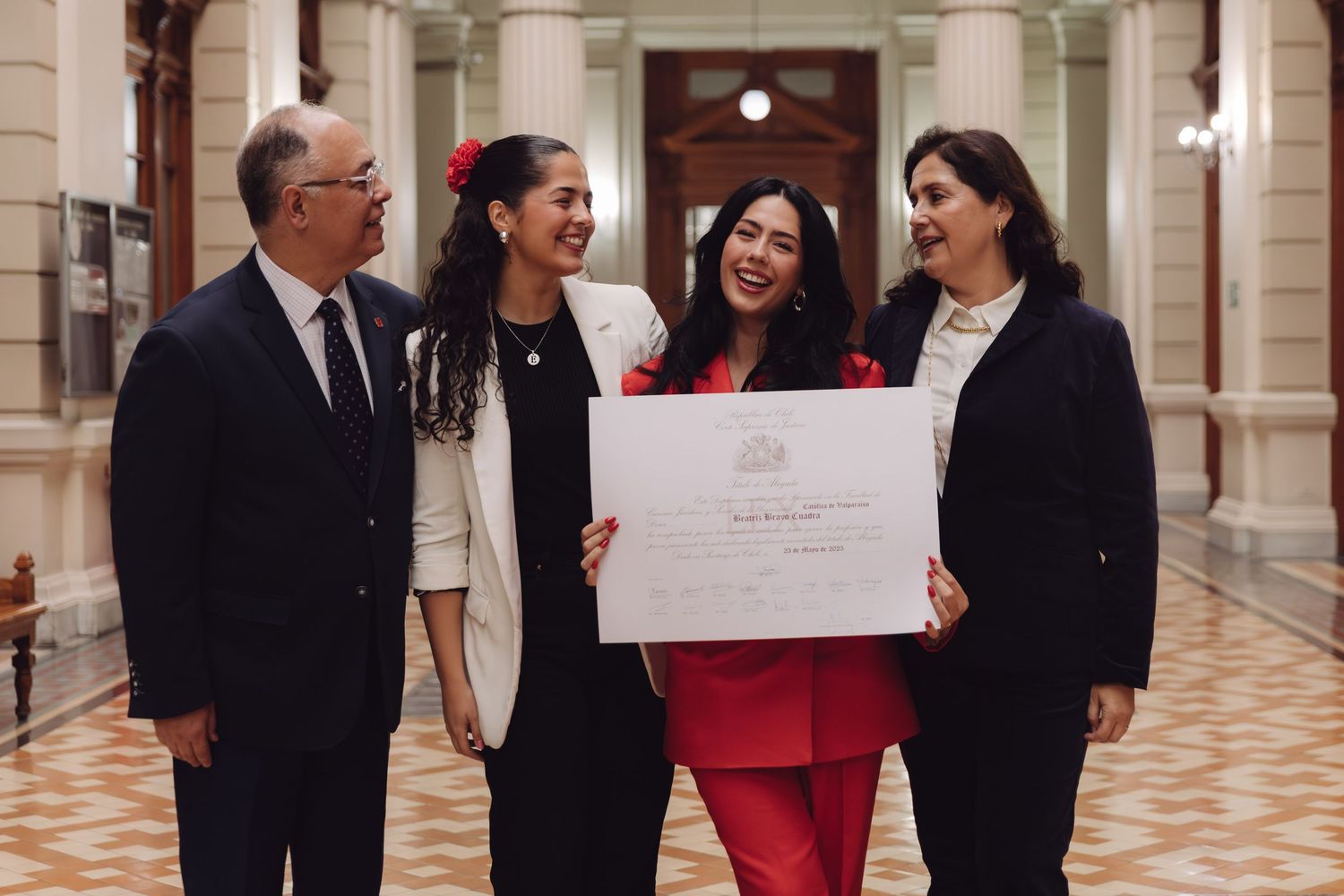 Laughing graduate in red suit celebrates with family holding diploma in a grand corridor.