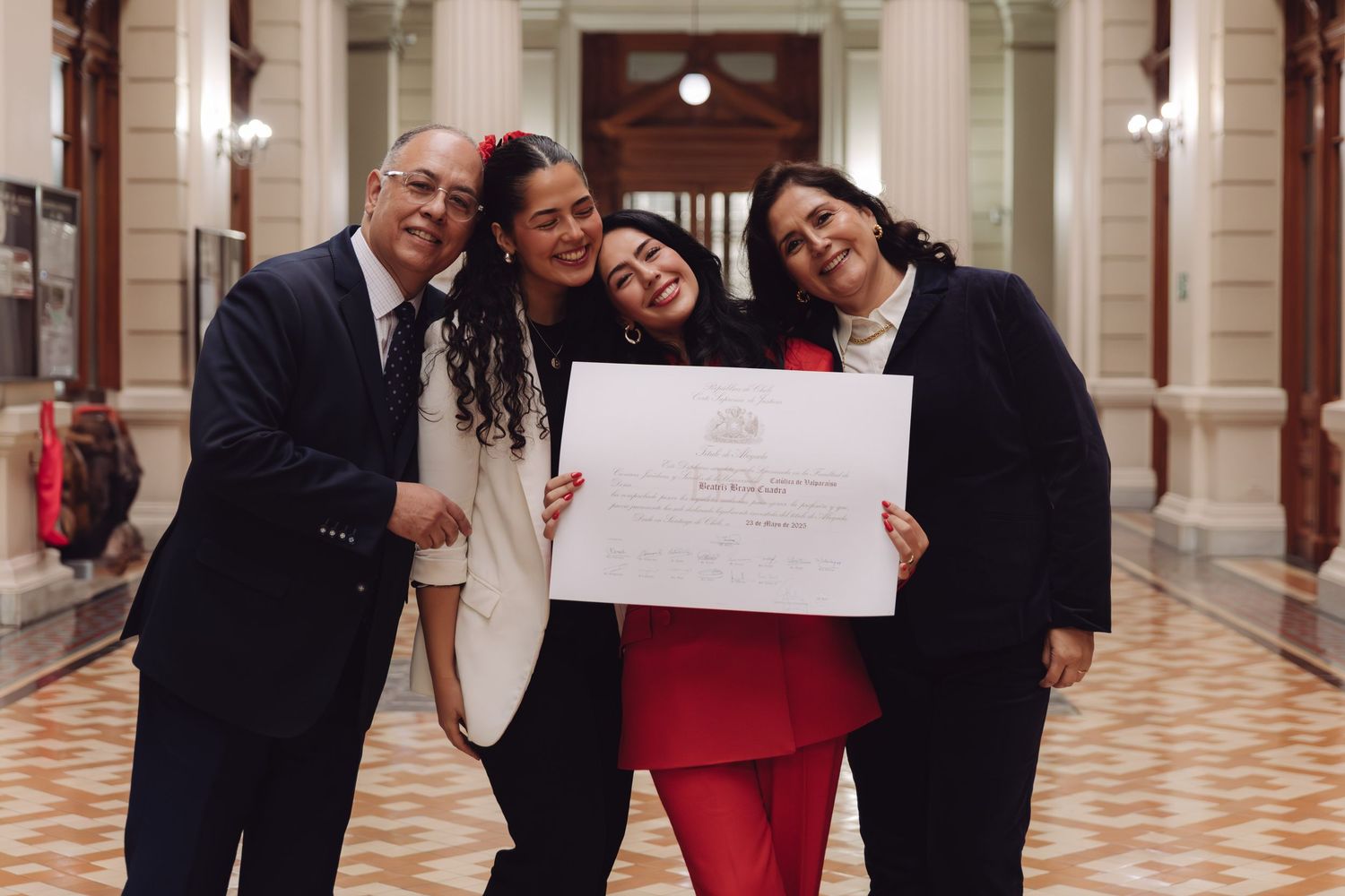 Graduate in red suit joyfully poses with family holding diploma in a classical building hallway.