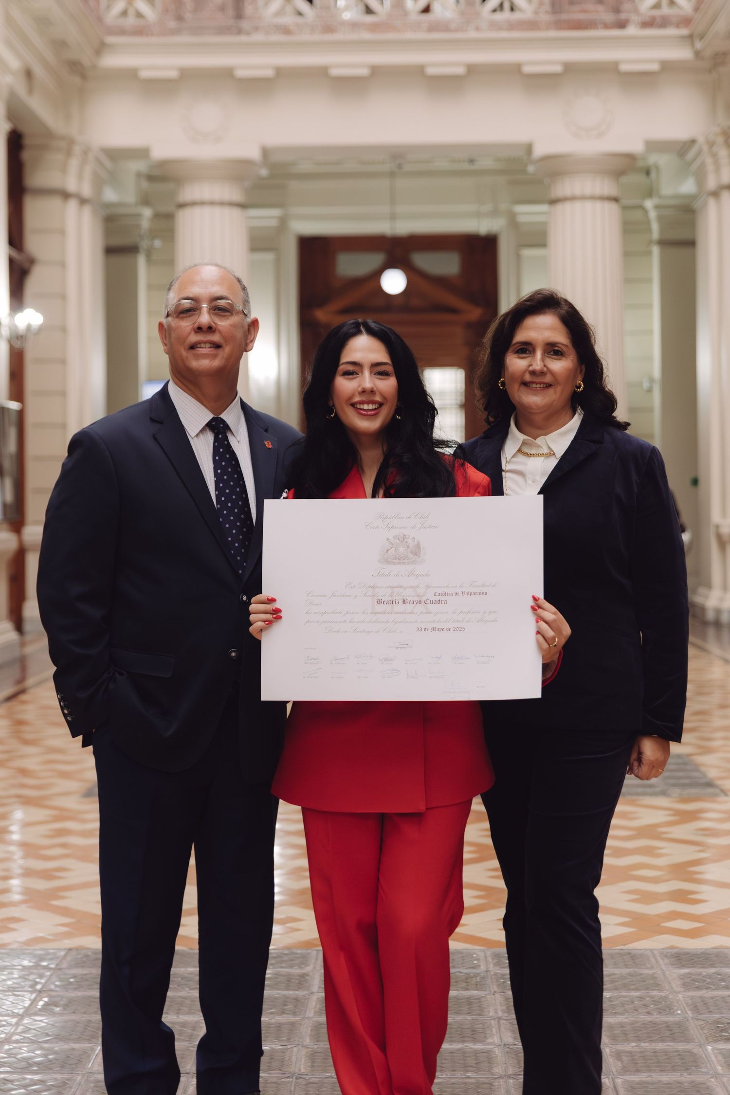 Three people pose with a diploma in an elegant columned hallway, woman in red suit flanked by two others.