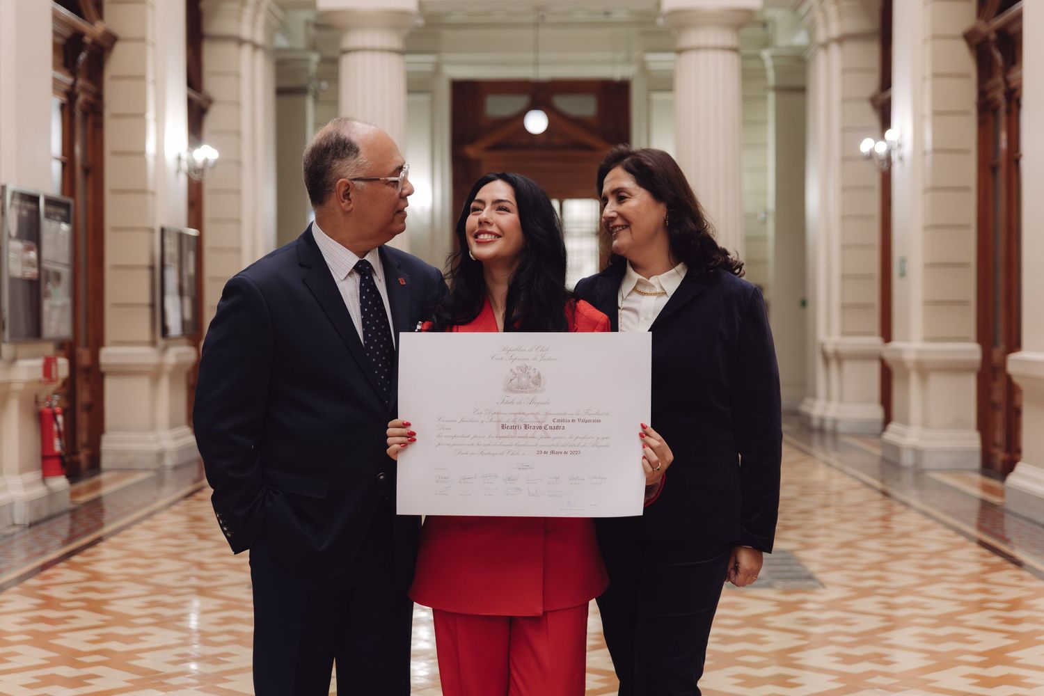 Three people laughing together holding a certificate in a grand classical building corridor.