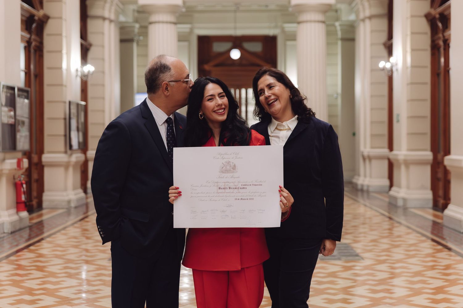 Man kisses woman in red suit holding diploma while another woman smiles in ornate hallway.