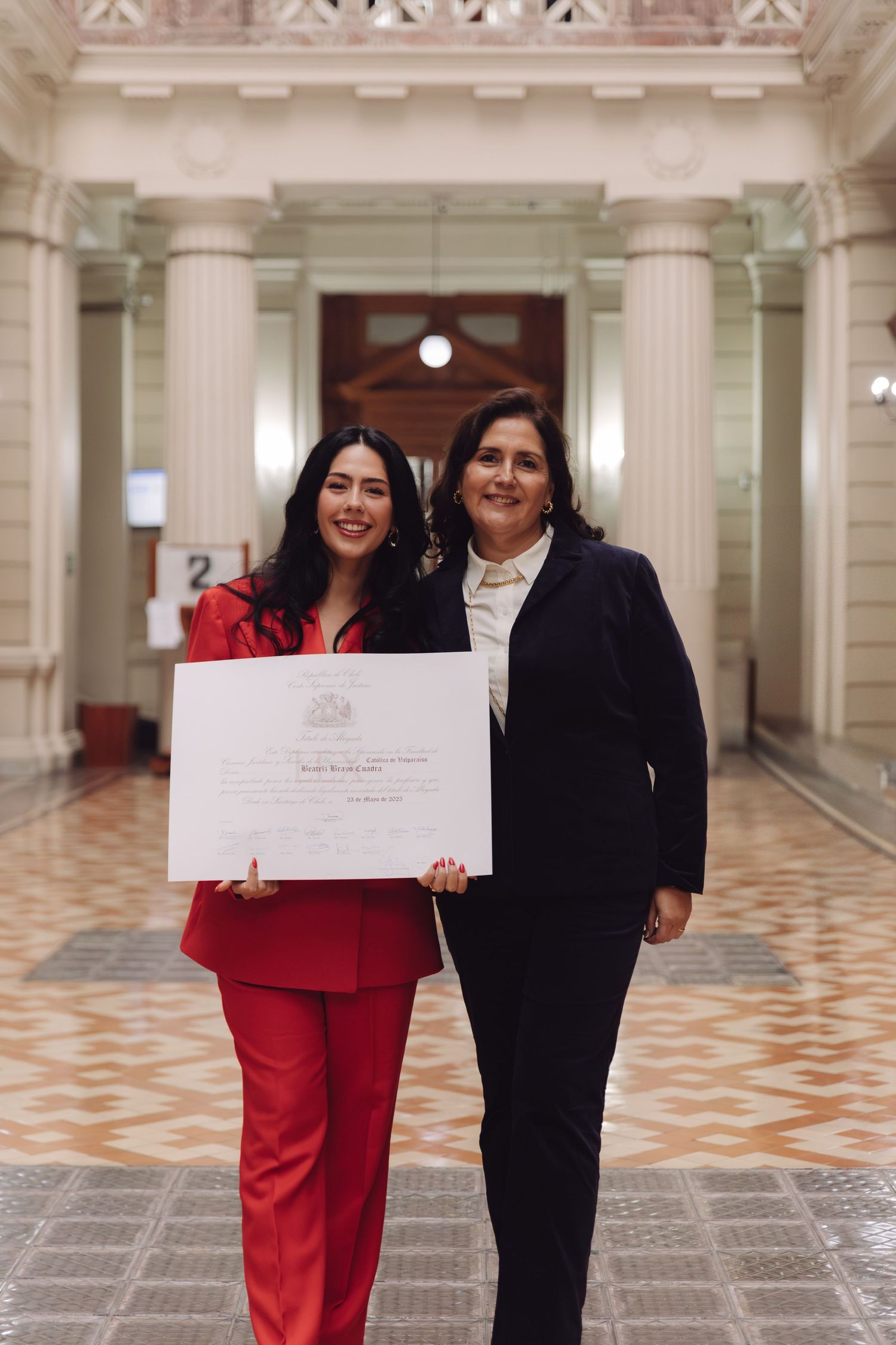 Two women pose with a graduation diploma in a neoclassical building corridor, one in red suit.