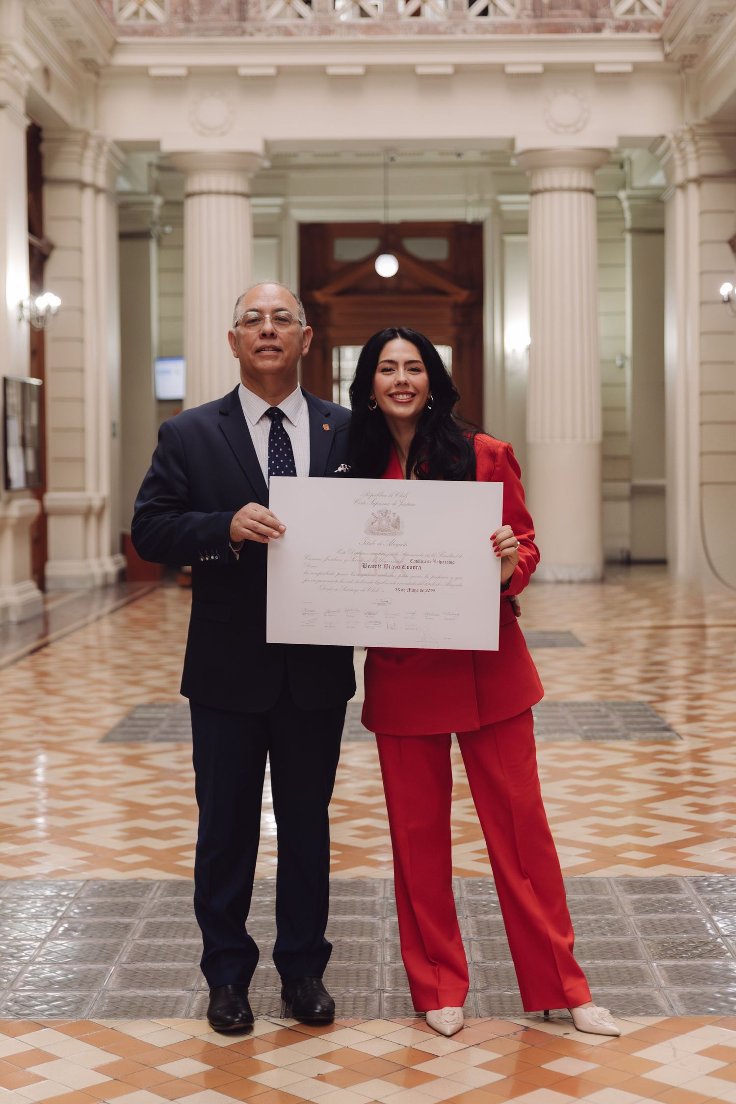 Man and woman in red suit proudly display official certificate in elegant columned hallway.