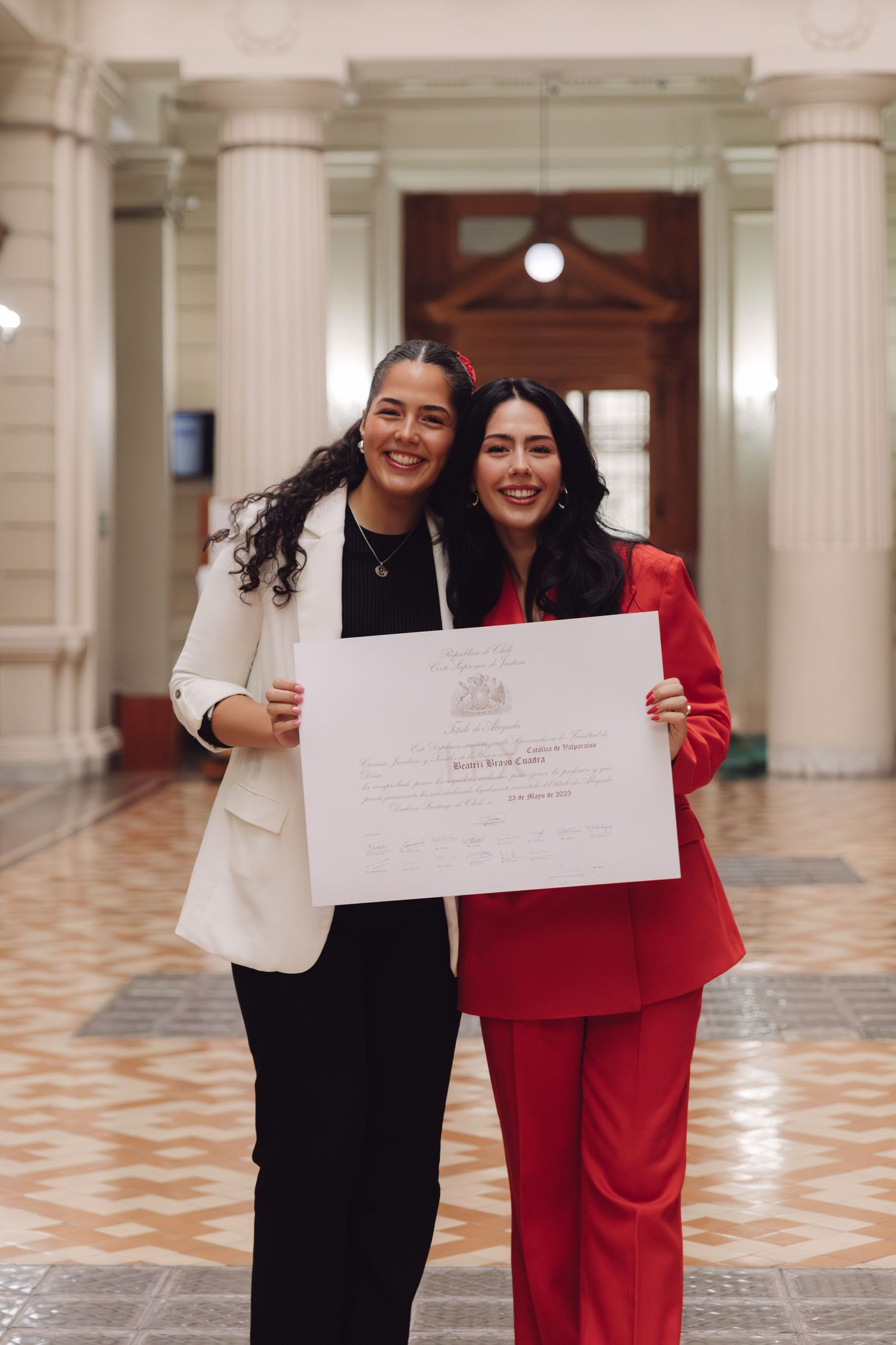 Two young women smile holding a large diploma certificate in a grand classical building hallway.