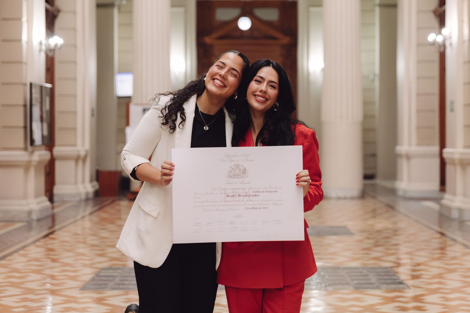 Two women laugh joyfully while holding a diploma in a beautiful columned corridor.