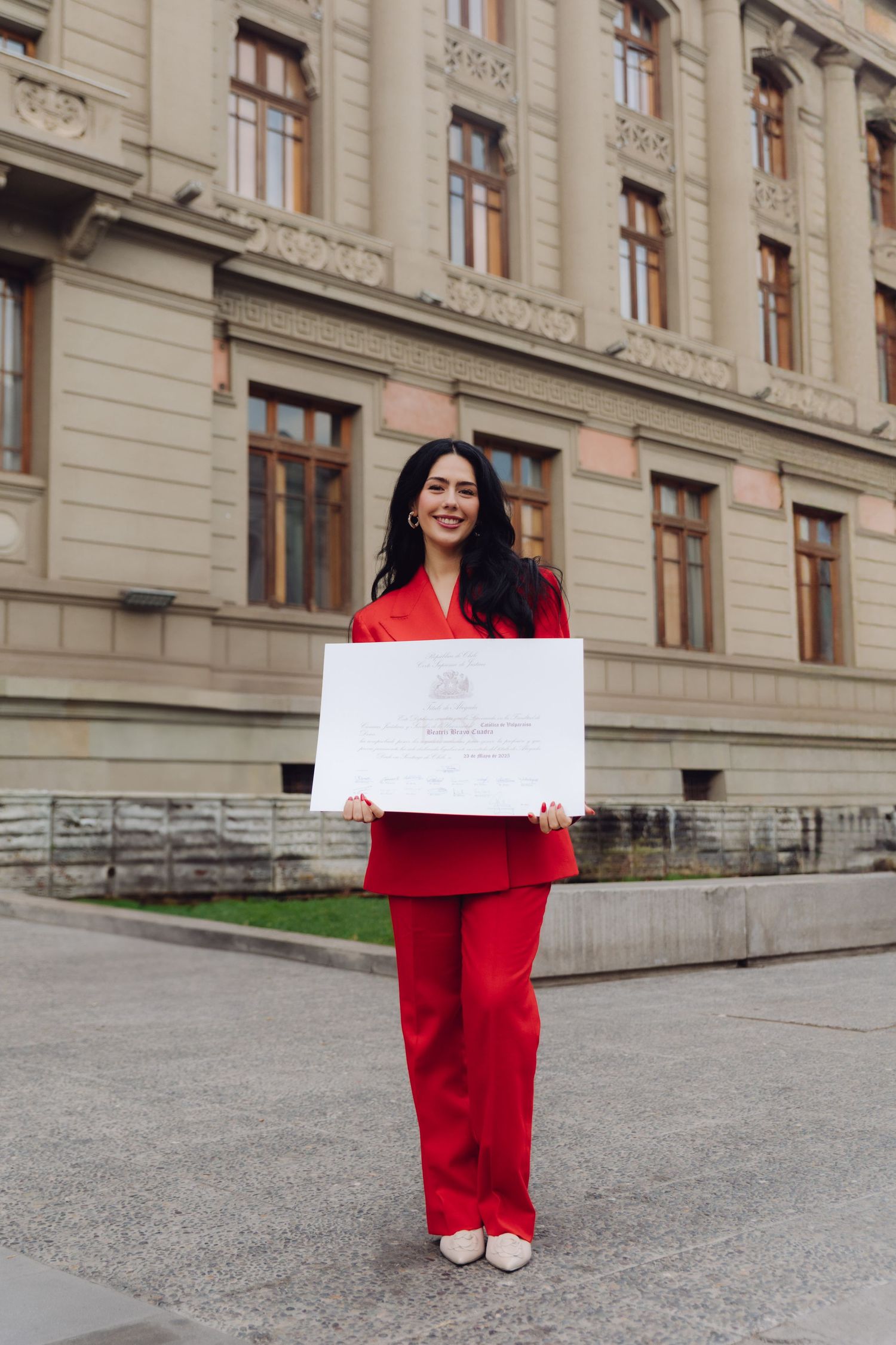 Woman in red suit proudly displaying a certificate outside a historic building.