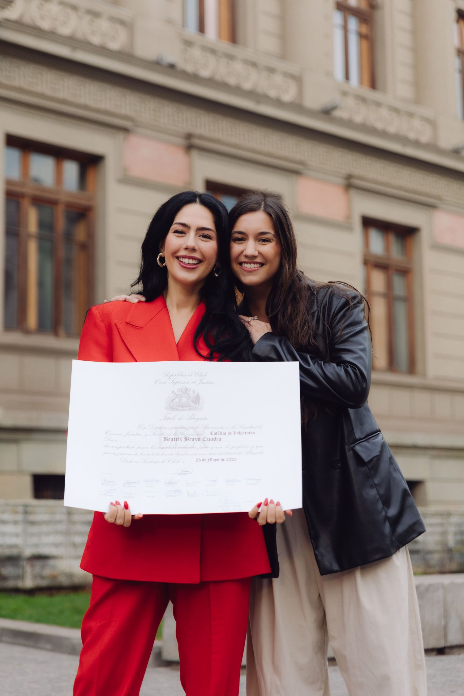Two women smiling and holding a certificate outside a European university building.