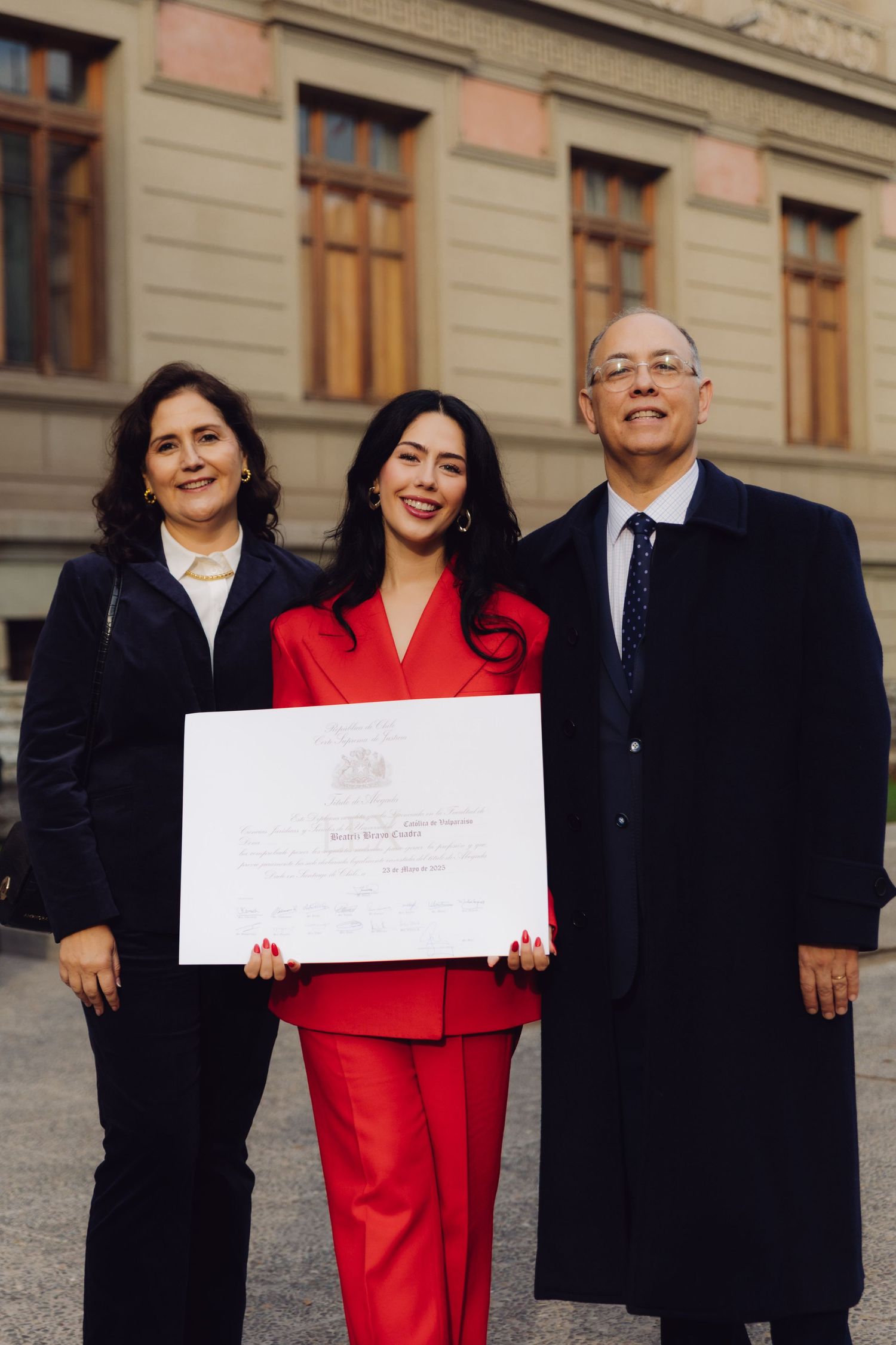 Woman in red suit holding a certificate flanked by her parents outside a building.