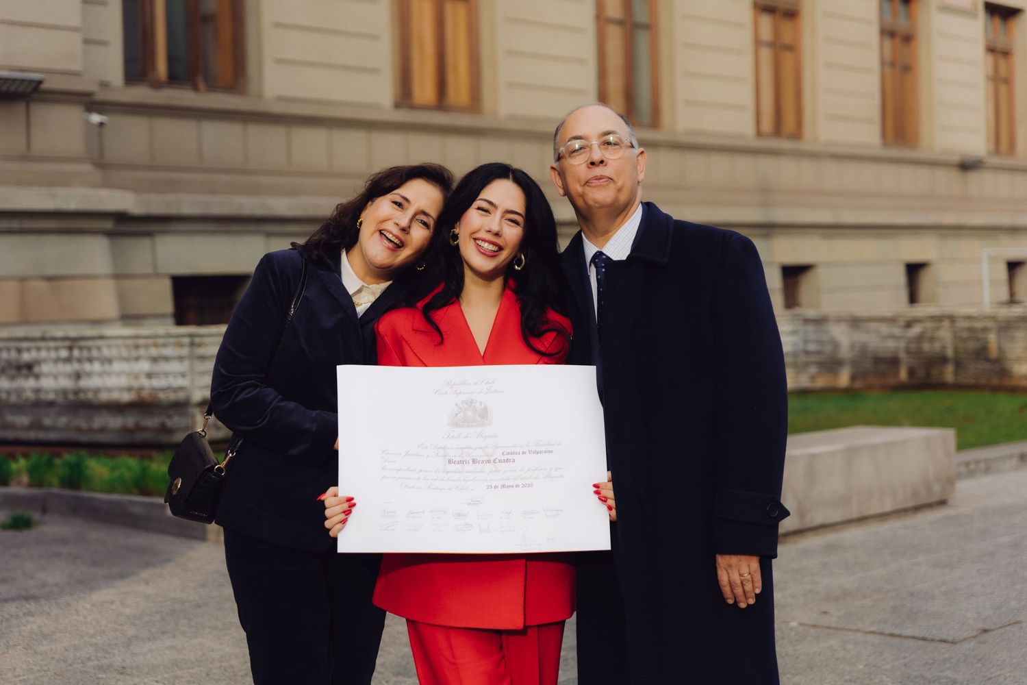 Family of three proudly displaying a certificate outside a classical building.